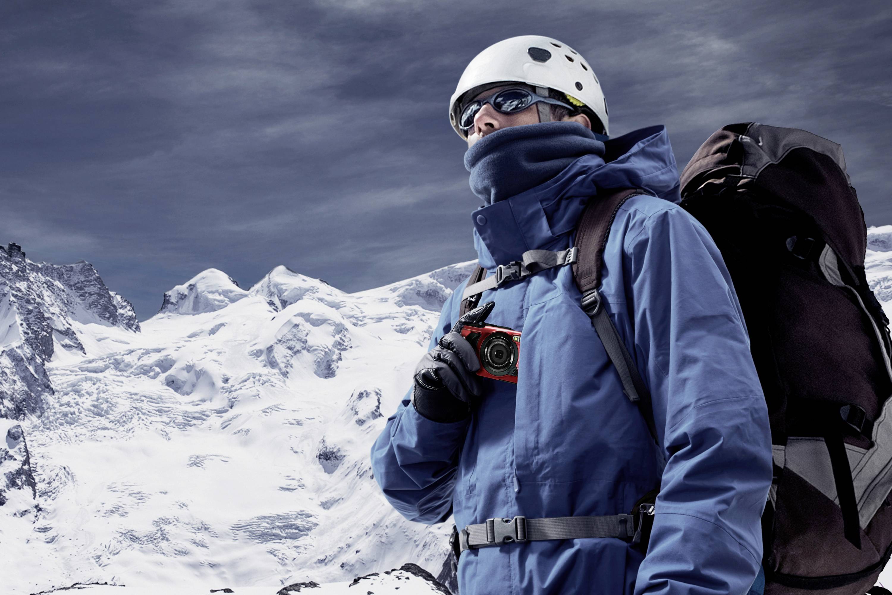 Alpinista w ciepłym wyposażeniu stoi przed ośnieżonymi górami, trzyma kompas i niesie duży plecak.