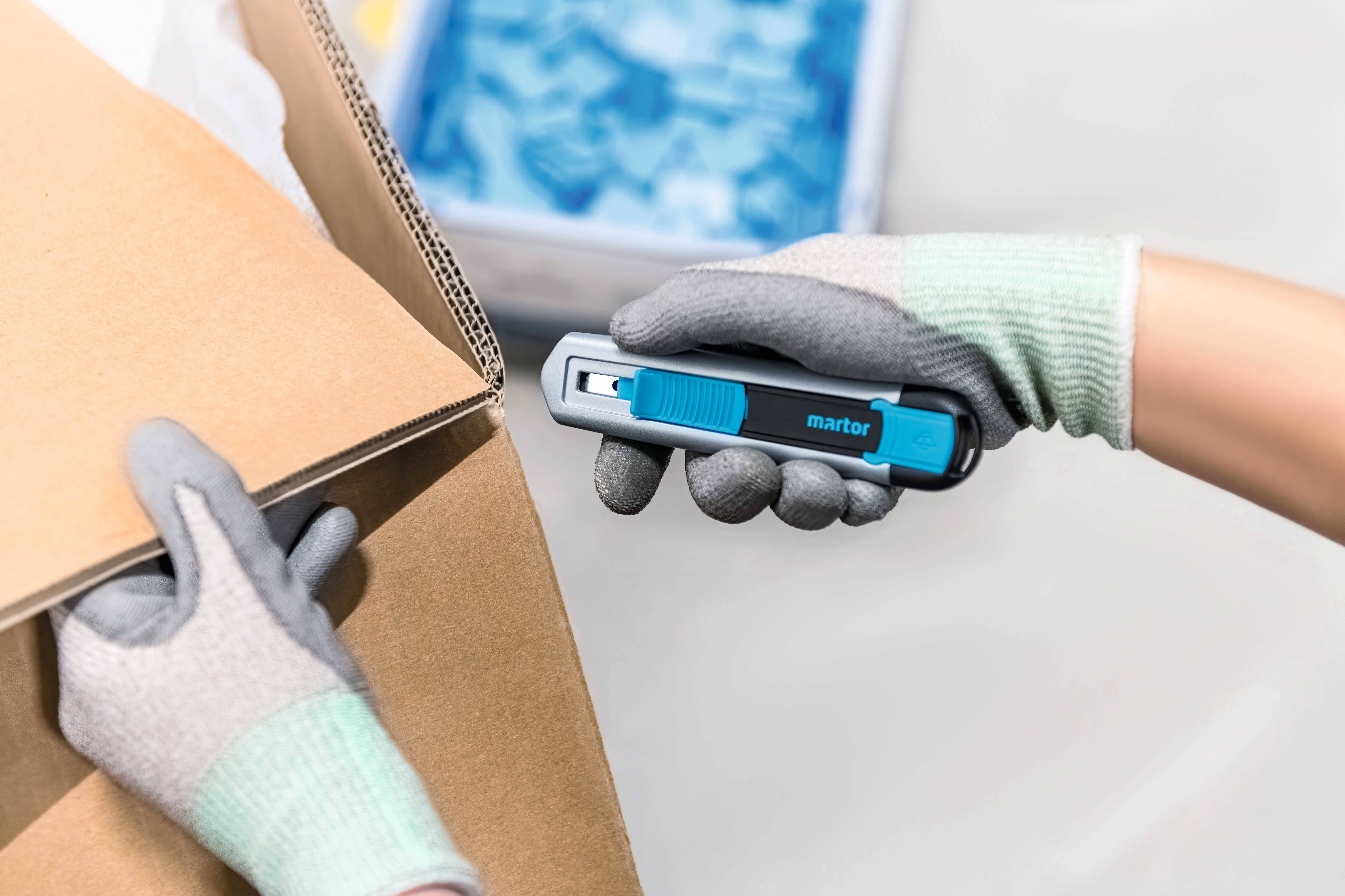 Osoba w rękawiczkach przecina nożem bezpieczeństwa karton. W tle widoczne są niebieskie obiekty.