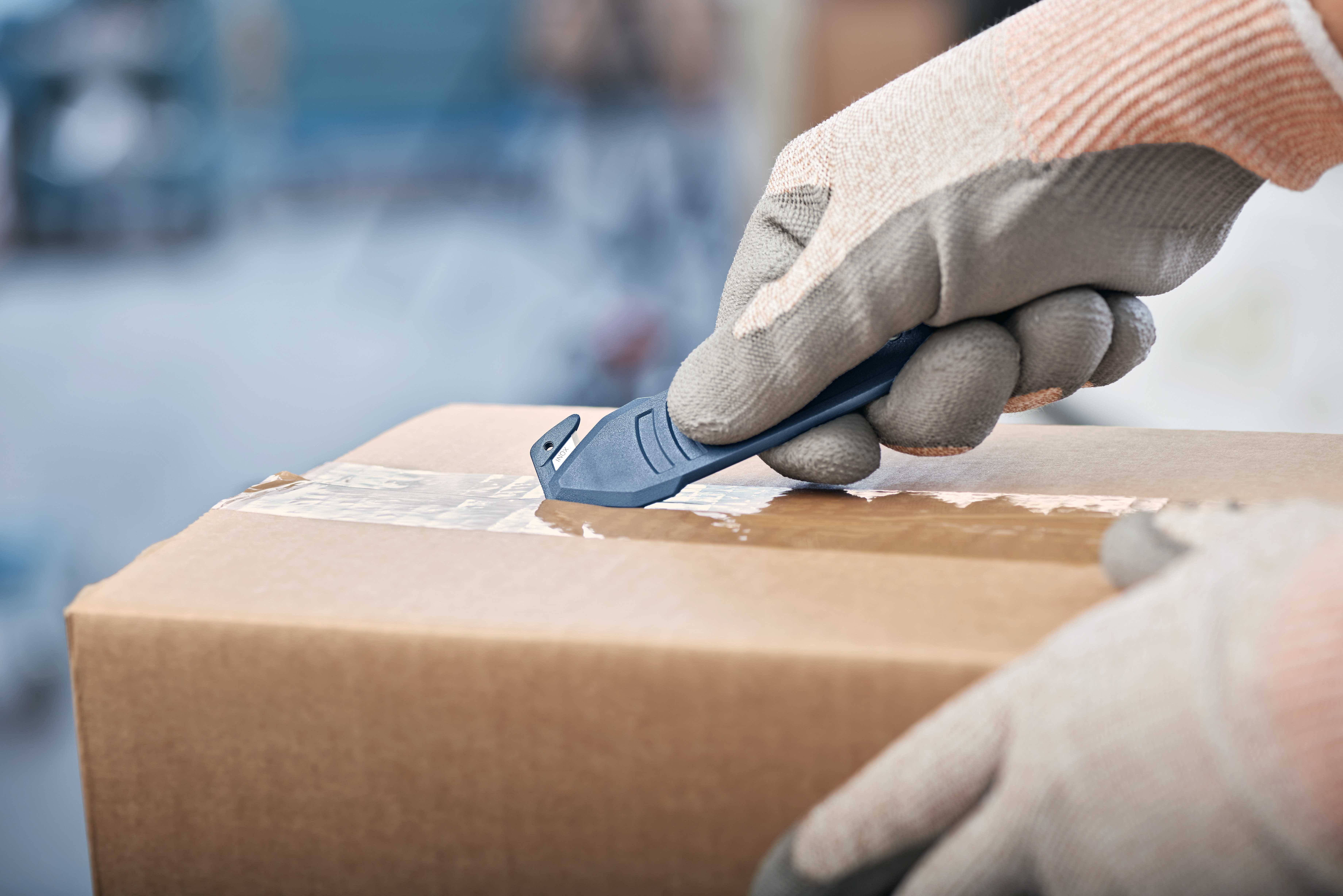 Osoba w rękawicach roboczych otwiera za pomocą noża zapieczętowany pakiet. Fokus spoczywa na precyzyjnym ruchu cięcia.