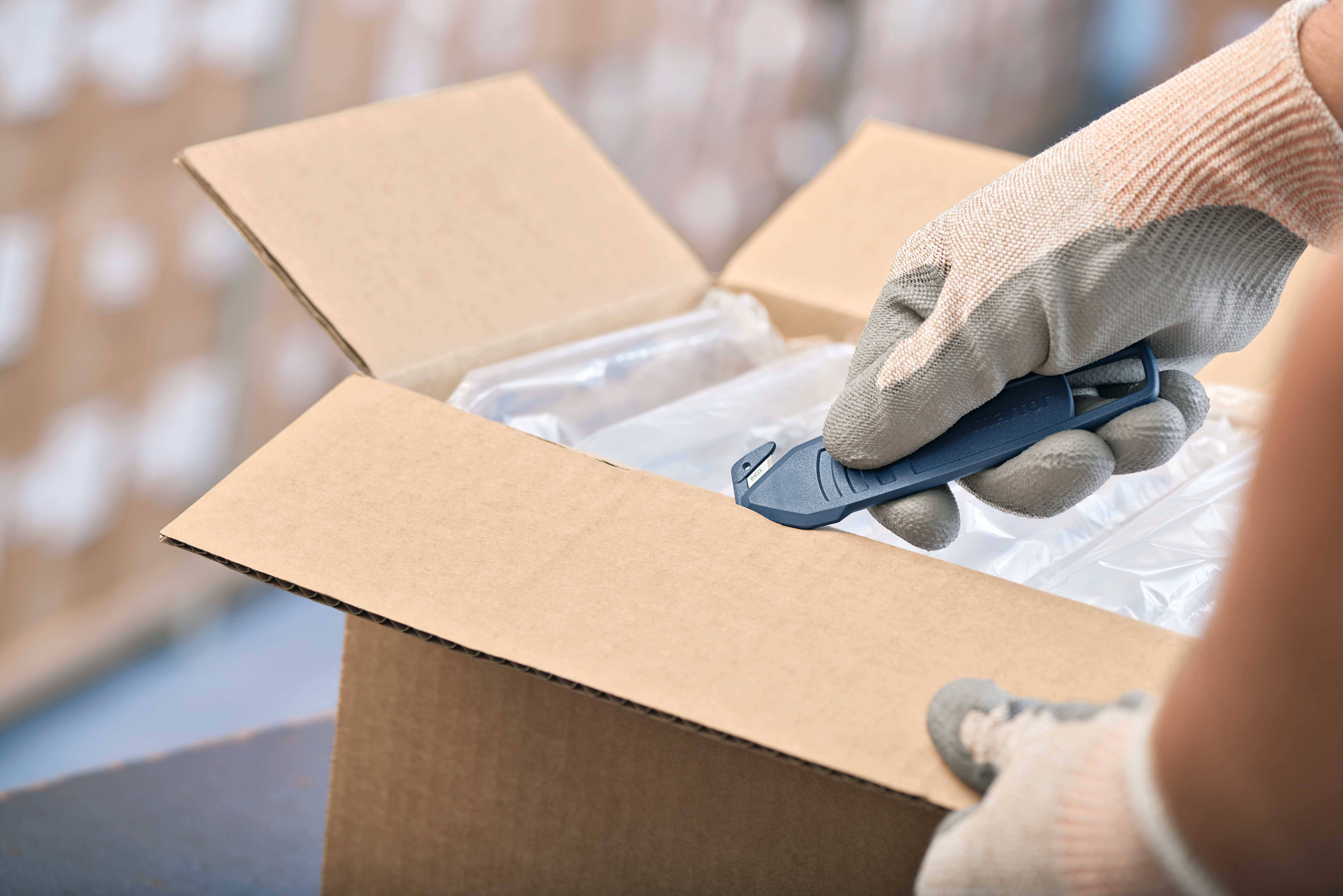 Dłoń w rękawicy otwiera karton za pomocą noża introligatorskiego. W tle widać rozmyte kartony w magazynie.