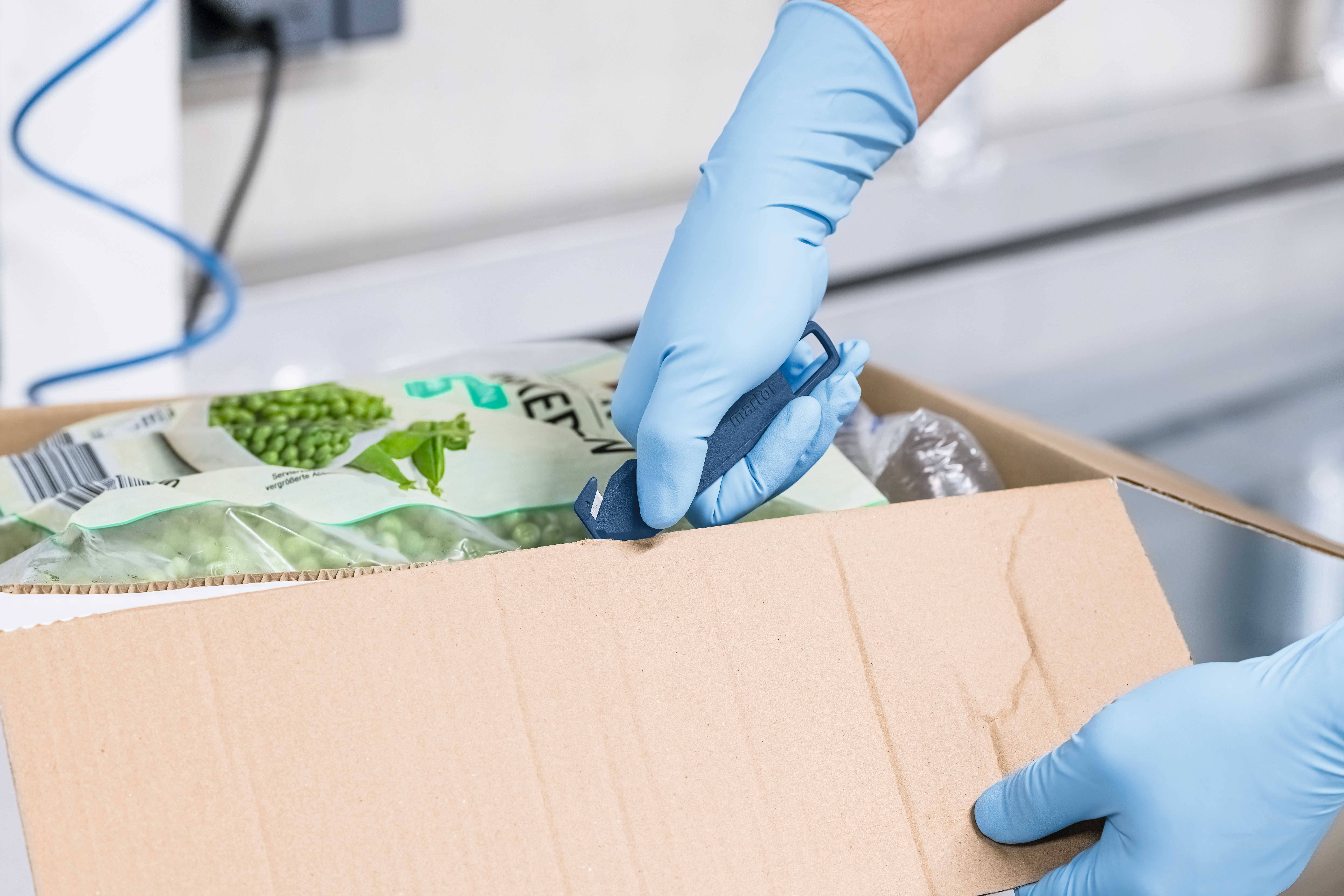 Osoba w niebieskich rękawiczkach otwiera kartonowe pudełko z mrożonym groszkiem.