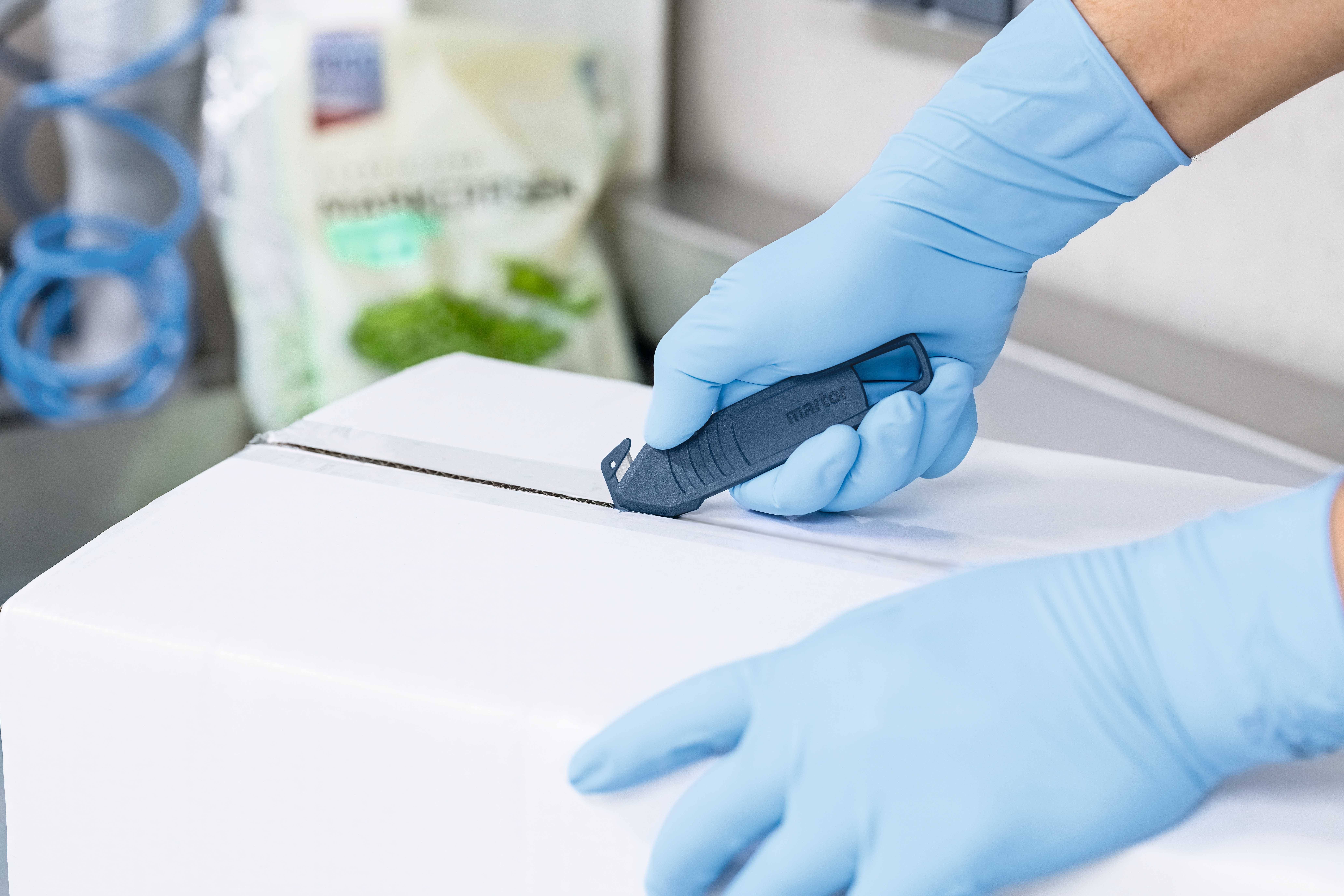 Osoba przecina karton nożem do tektury, używając niebieskich jednorazowych rękawic ochronnych. W tle rozmyty jest widoczny karton sałaty.