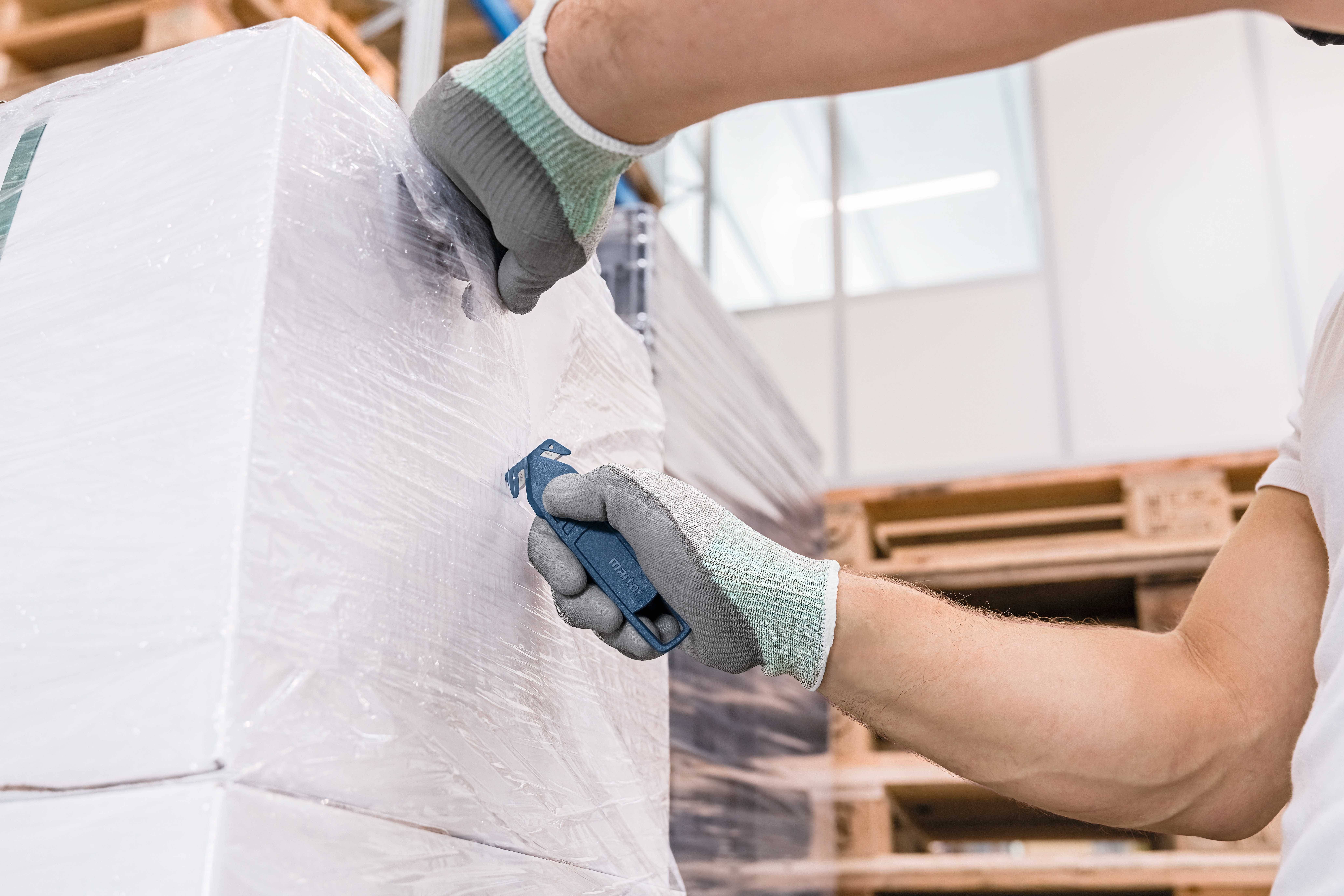 Osoba w rękawiczkach ucina folię plastikową z palety w magazynie. W tle widoczne są regały.