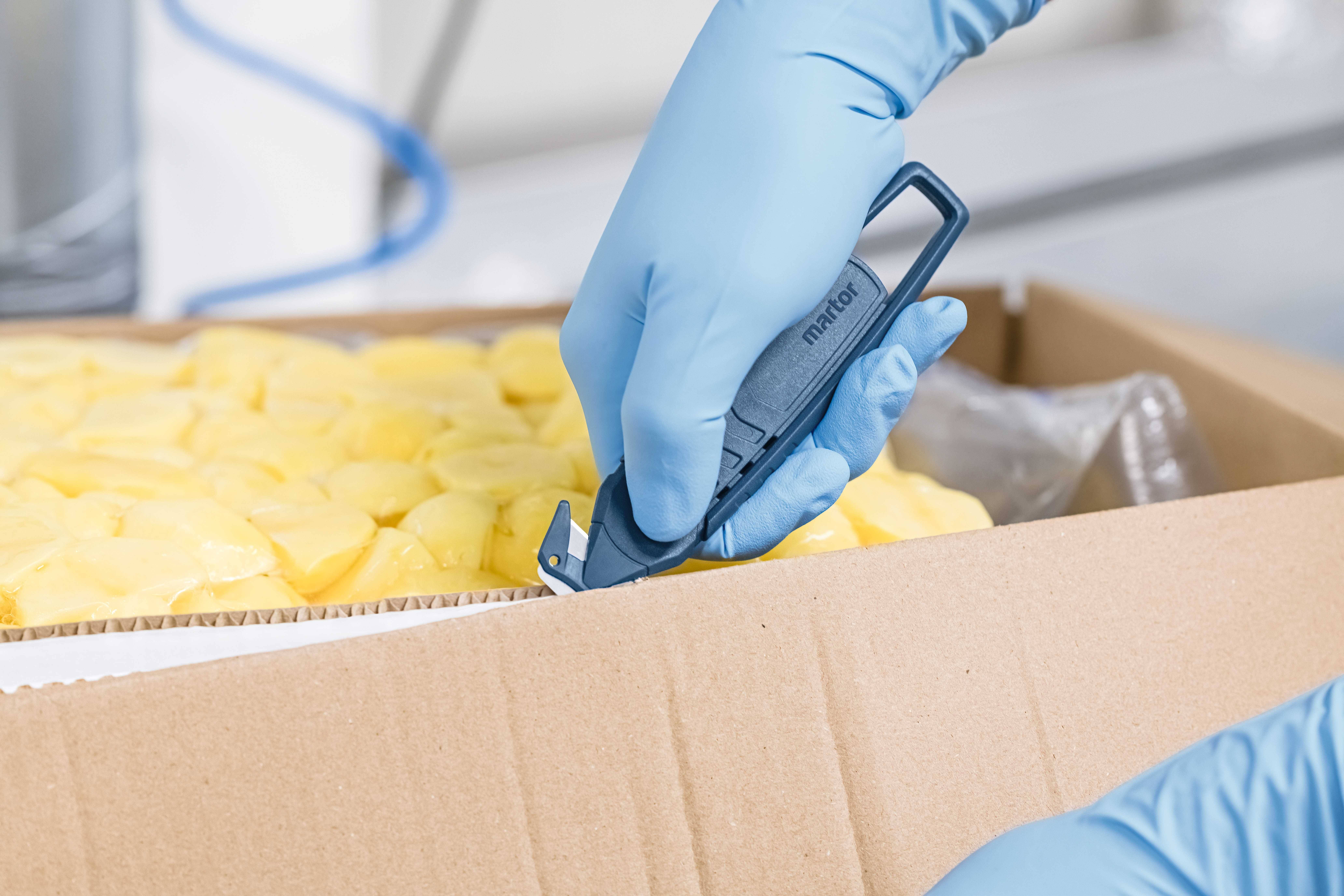 Ręka w niebieskiej rękawicy otwiera karton pełen żółtych produktów spożywczych za pomocą narzędzia tnącego.