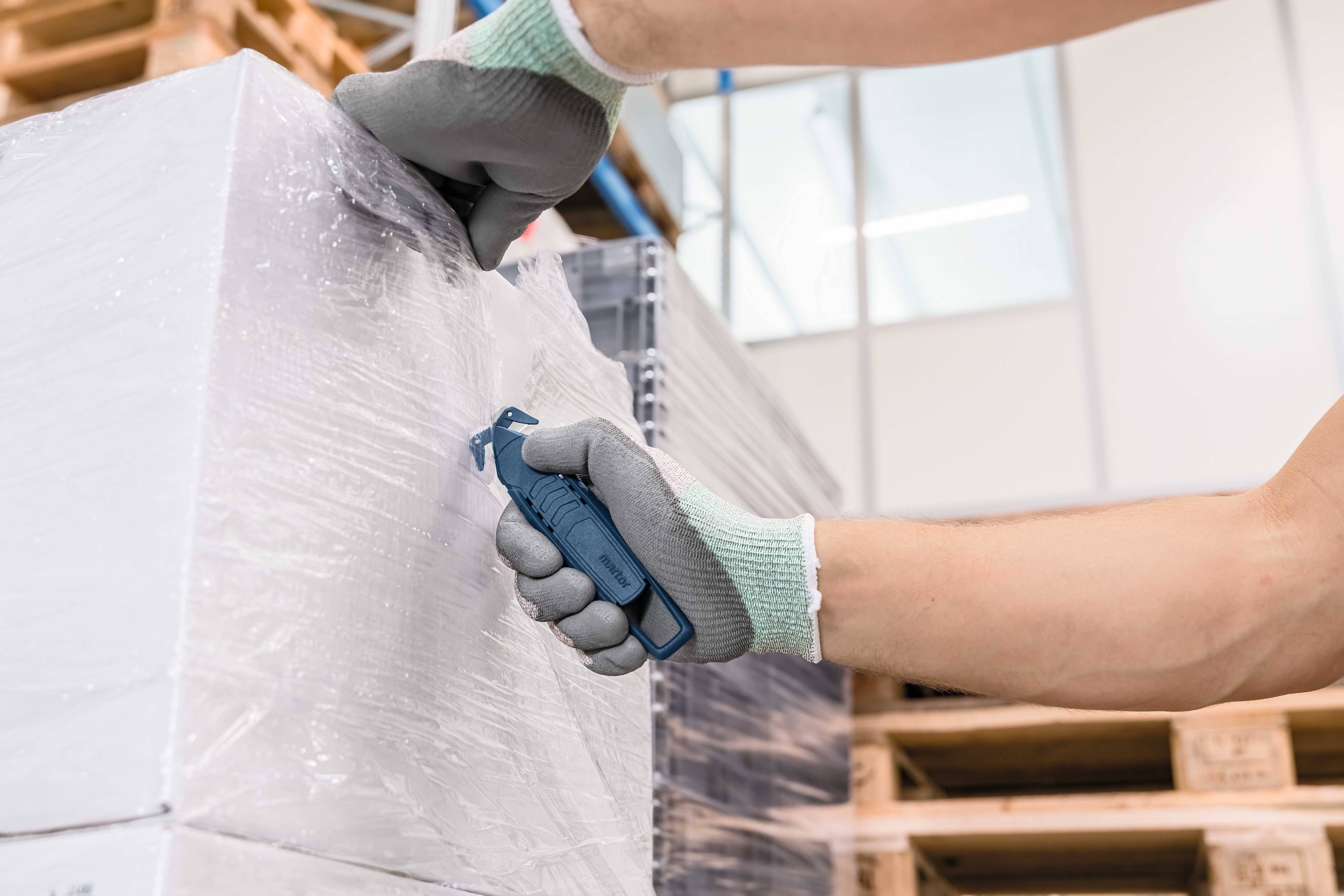 Osoba ucina folię plastikową z palety za pomocą noża bezpiecznego. Rękawice chronią jej dłonie.