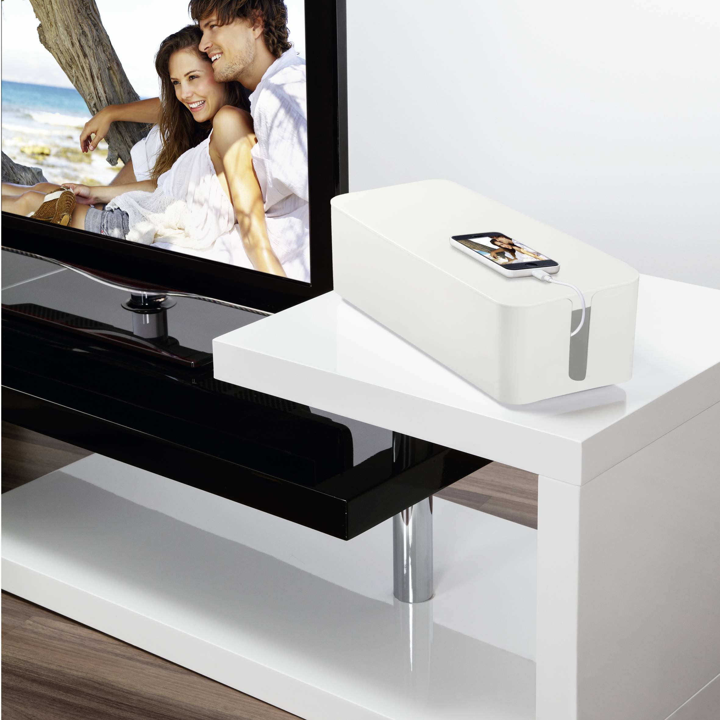 Biały stół z pudełkiem na kable, obok płaskiego telewizora wyświetlającego parę na plaży.