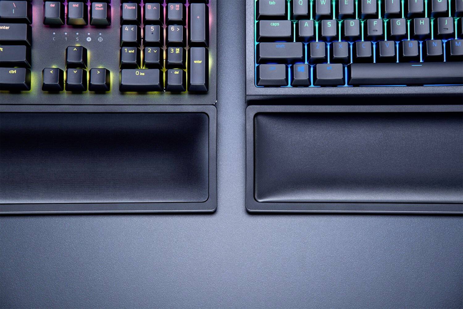 Dwie mechaniczne klawiatury z kolorowym podświetleniem w tle i podpórkami na nadgarstki na stole.