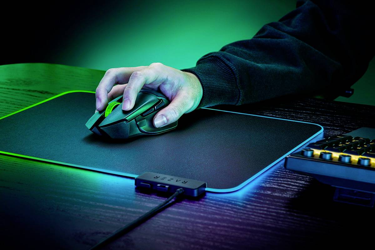 Osoba obsługuje mysz komputerową na świecącej podkładce RGB, obok której znajduje się klawiatura z żółtym podświetleniem w tle.