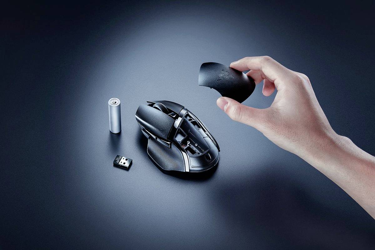 Dłoń kładzie górną część obudowy na myszce gamingowej, obok niej bateria i bezprzewodowy odbiornik USB. Nacisk na przyjazny użytkownikowi design i akcesoria.