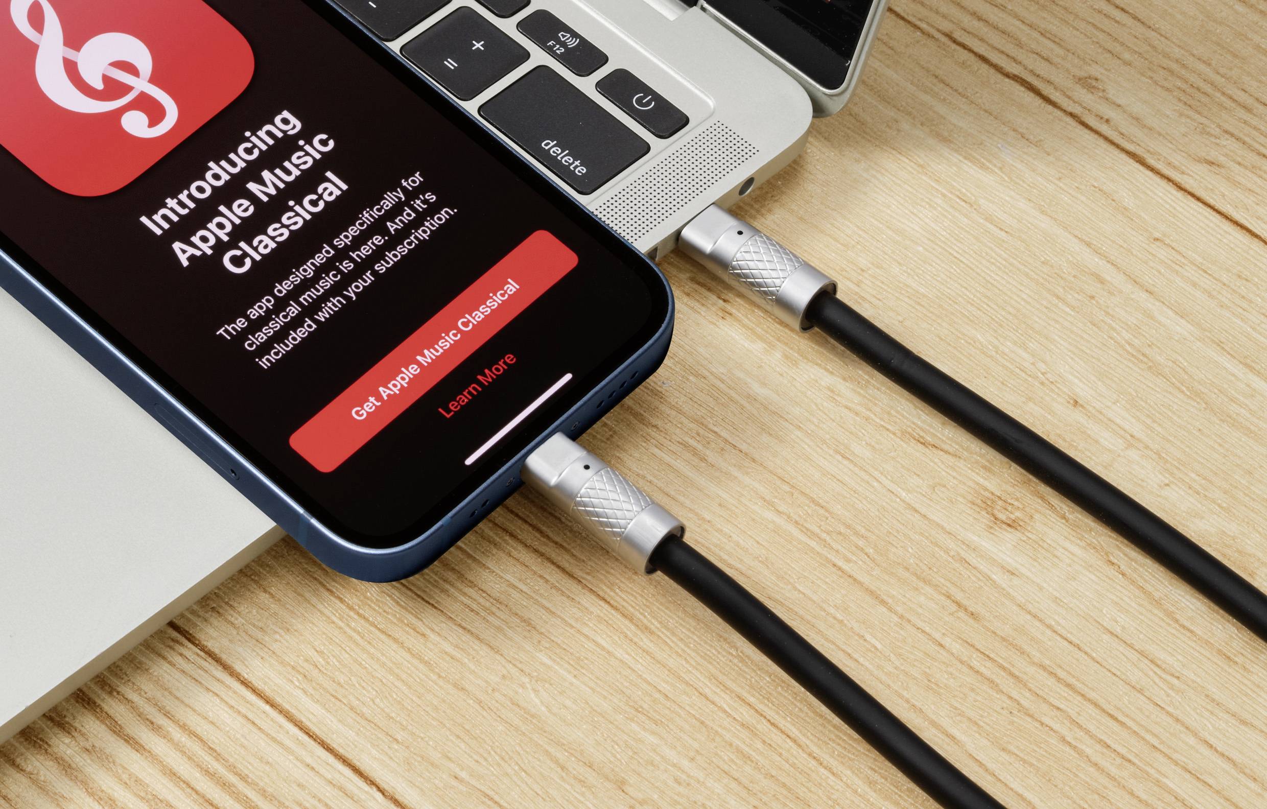Smartfon na laptopie z wyświetlaczem "Introducing Apple Music Classical", połączony dwoma kablami USB na drewnianym stole.