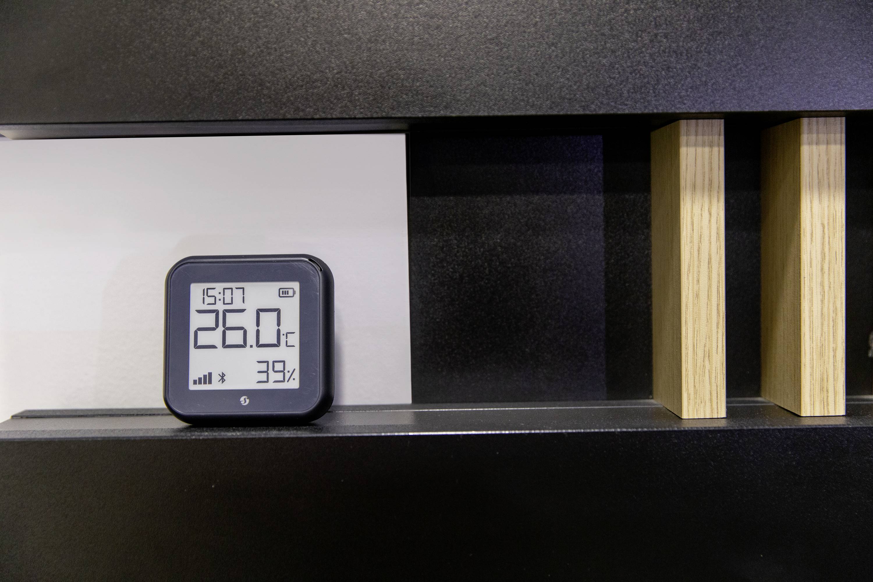 Cyfrowy termometr na ścianie wskazuje 26,0°C i 39% wilgotności. Godzina 15:07. Czarne tło z drewnianymi detalami.