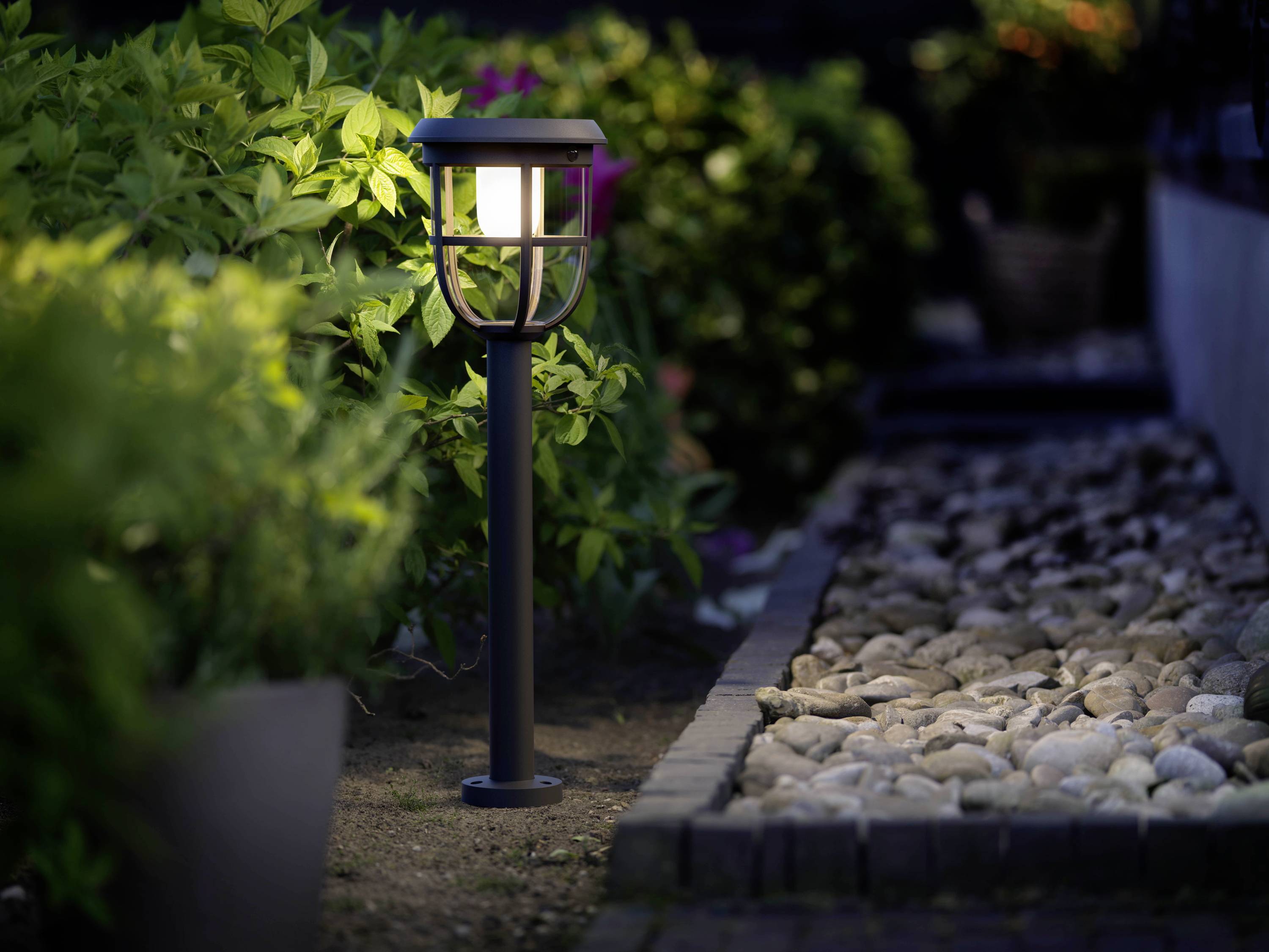 Lampa ogrodowa świeci w zmierzchu, otoczona roślinami i kamienistą ścieżką biegnącą po prawej stronie.