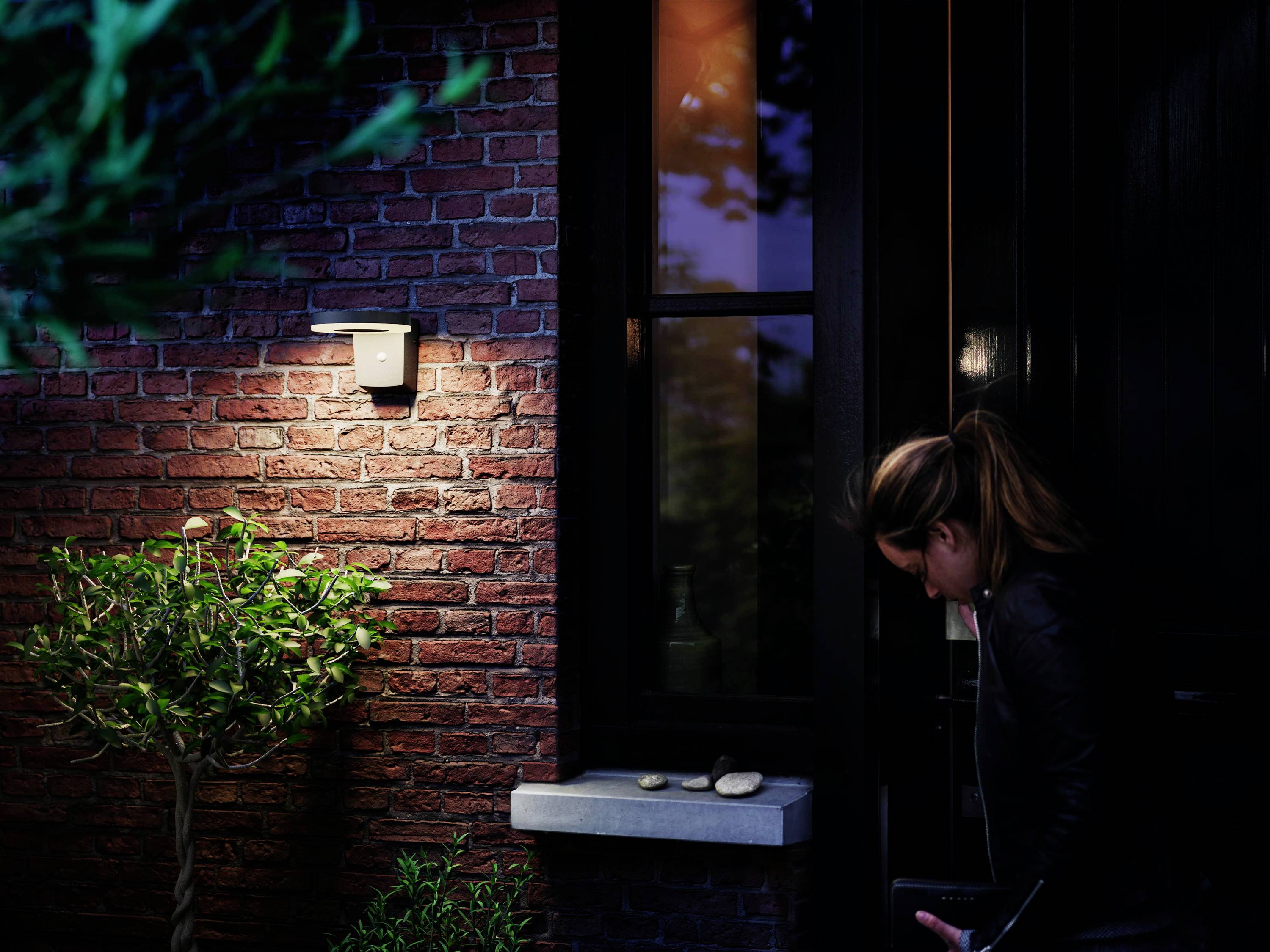 Kobieta w ciemności otwiera drzwi wejściowe domu z czerwonej cegły, oświetlona przez lampę zewnętrzną. Obok stoi mały drzewka.