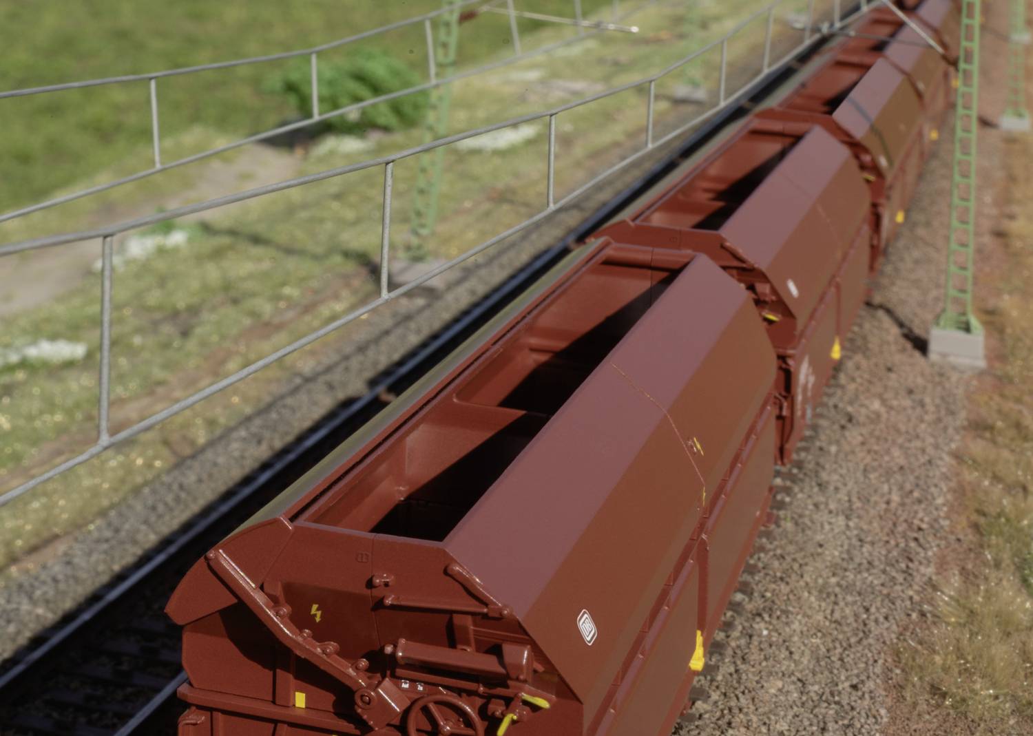 Pociąg towarowy z brązowymi wagonami jedzie po torach. Otoczenie składa się z trawy i ogrodzenia elektrycznego. Sfotografowany ukośnie z góry.