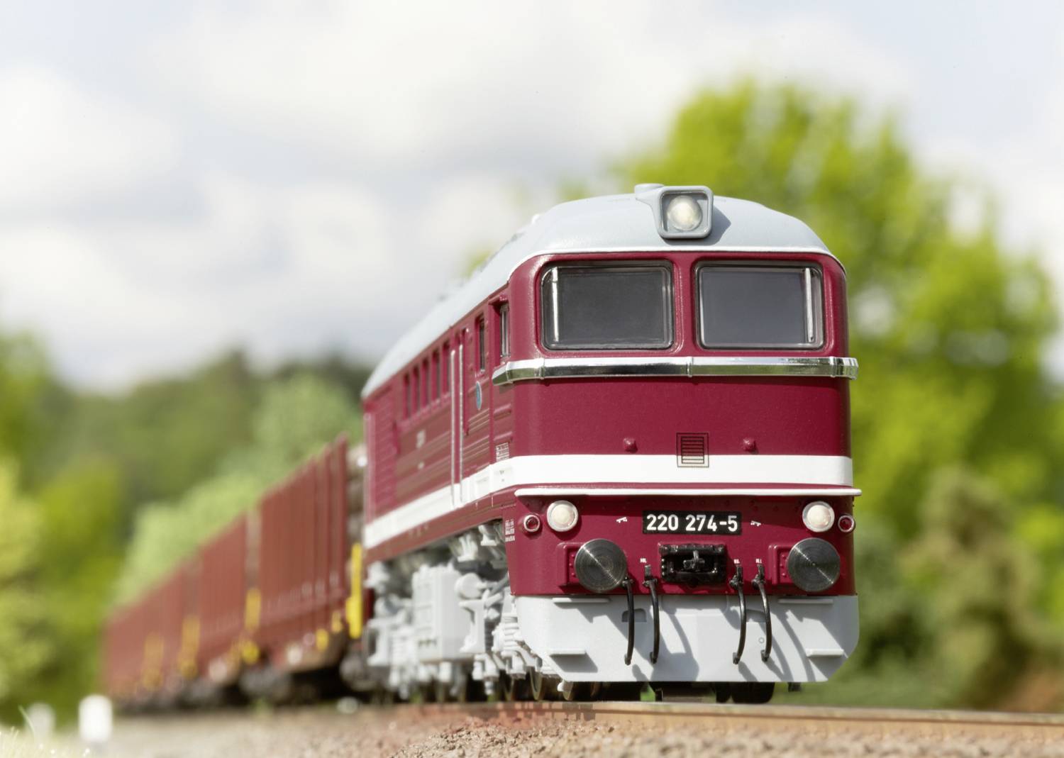 Czerwony pociąg towarowy jedzie po torach przez zieloną scenerię. Na pierwszym planie widoczna jest lokomotywa o numerze '220 274-5'.