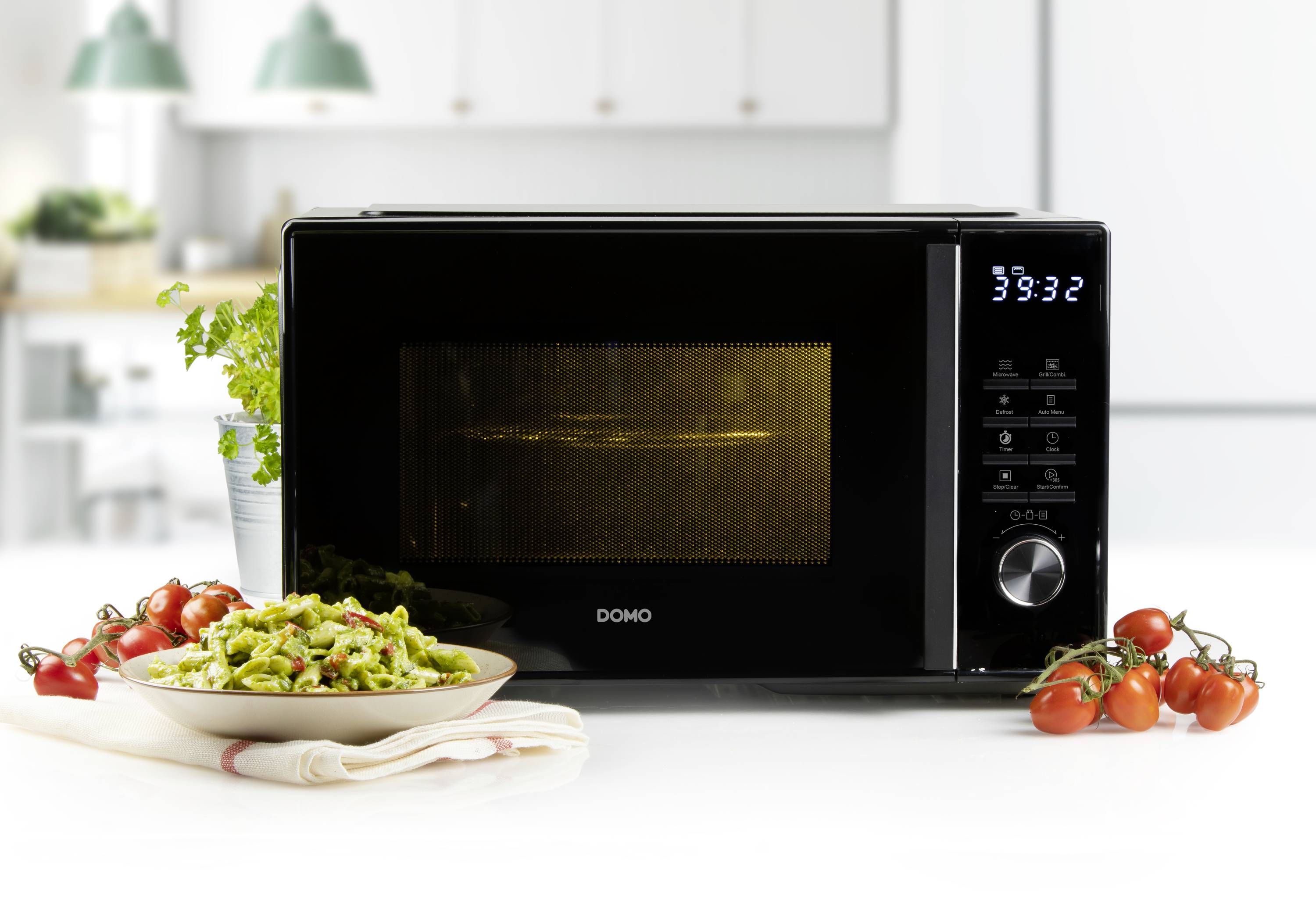 Czarna mikrofalówka stoi na białym stole w kuchni. Obok leży talerz z zielonym makaronem i pomidorkami koktajlowymi.