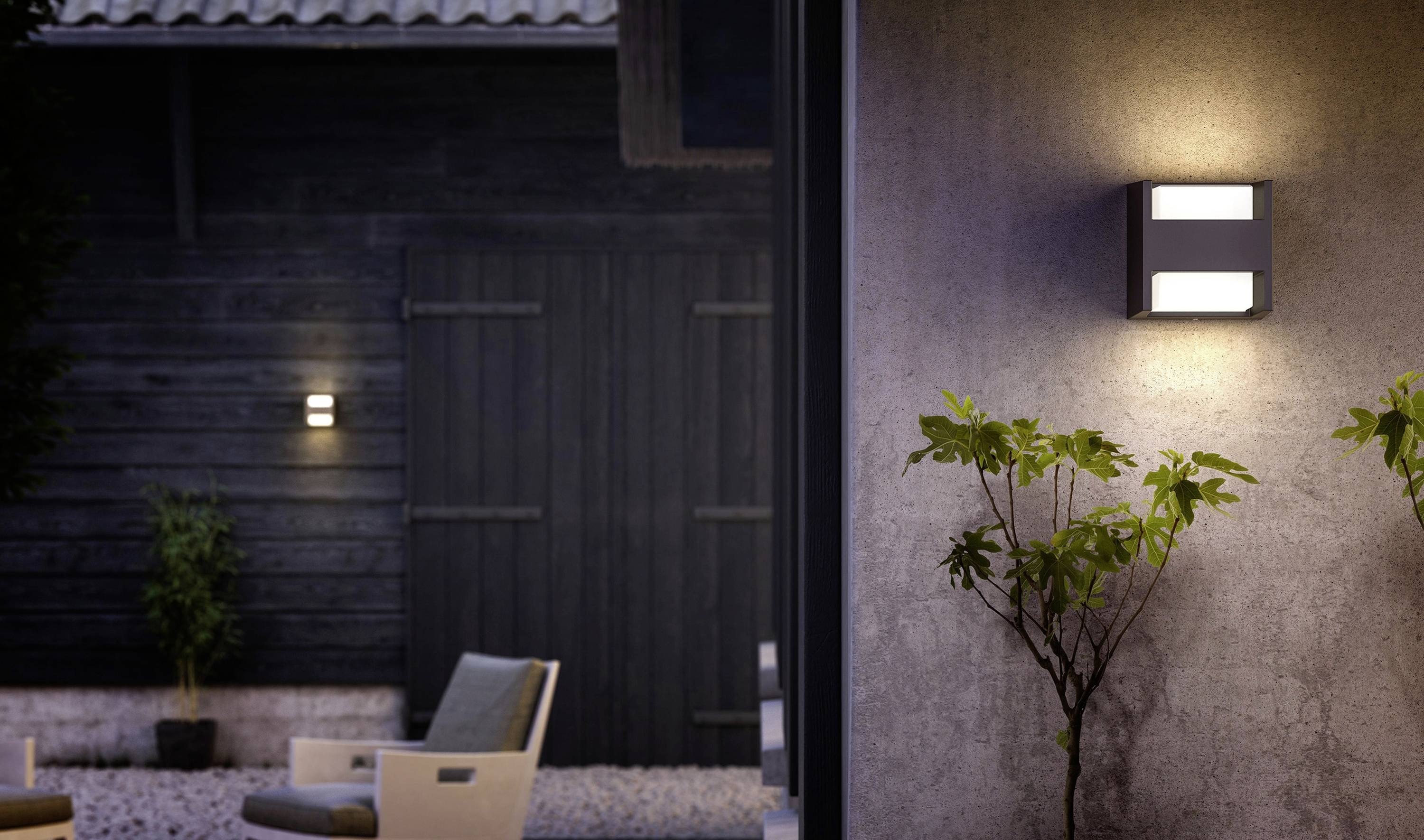 Nowoczesna lampa ścienna oświetla taras z dwoma krzesłami. W tle świeci kolejne światło na ciemnej drewnianej elewacji.