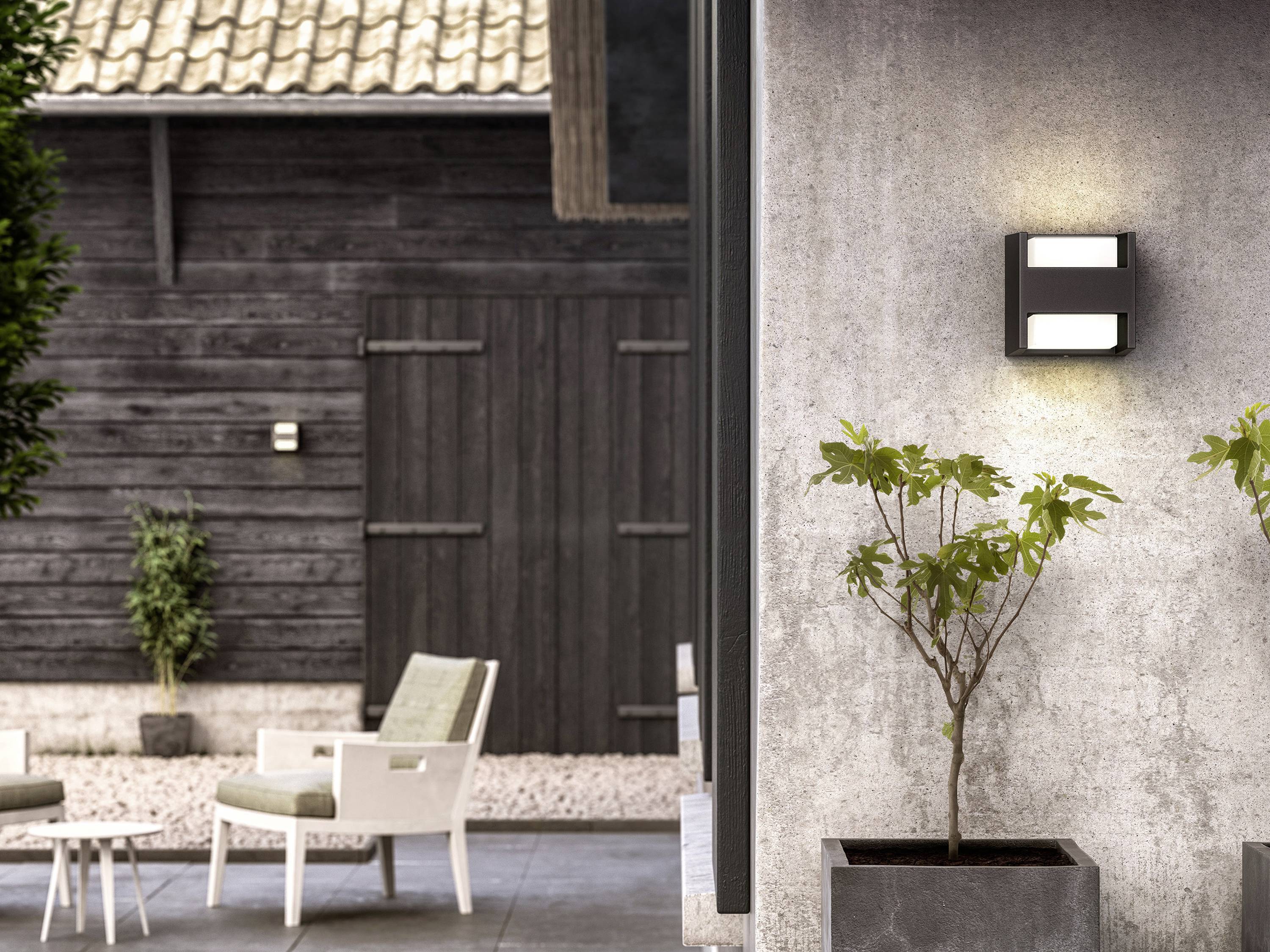 Nowoczesna przestrzeń zewnętrzna z drewnianymi krzesłami, doniczkowymi roślinami oraz podświetloną lampą ścienną przy betonowym domu. Prosta elegancja w ogrodzie.