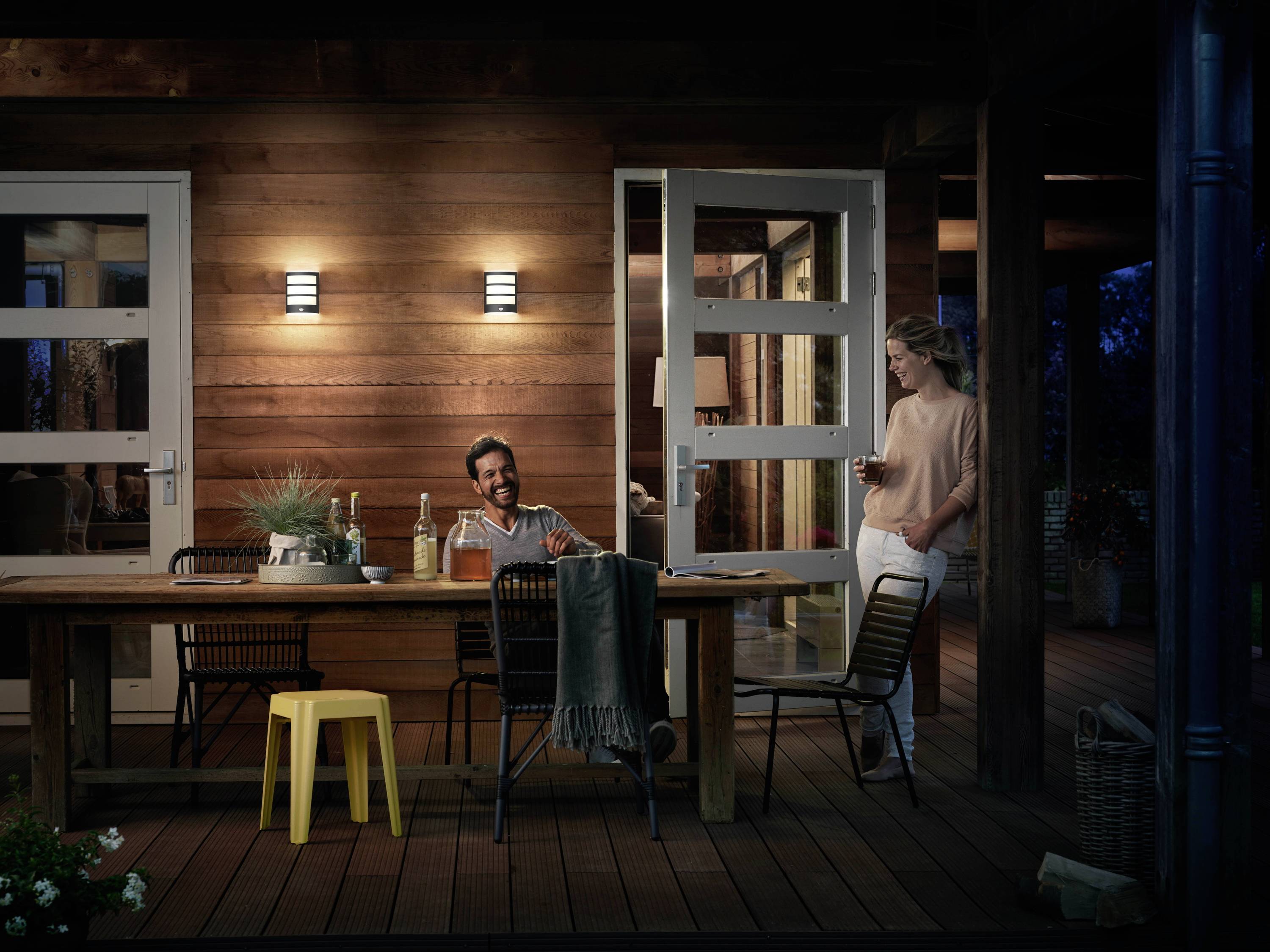 Kobieta i mężczyzna siedzą wieczorem na oświetlonej drewnianej tarasie. Mężczyzna siedzi, a kobieta stoi, trzymając w ręku napój.