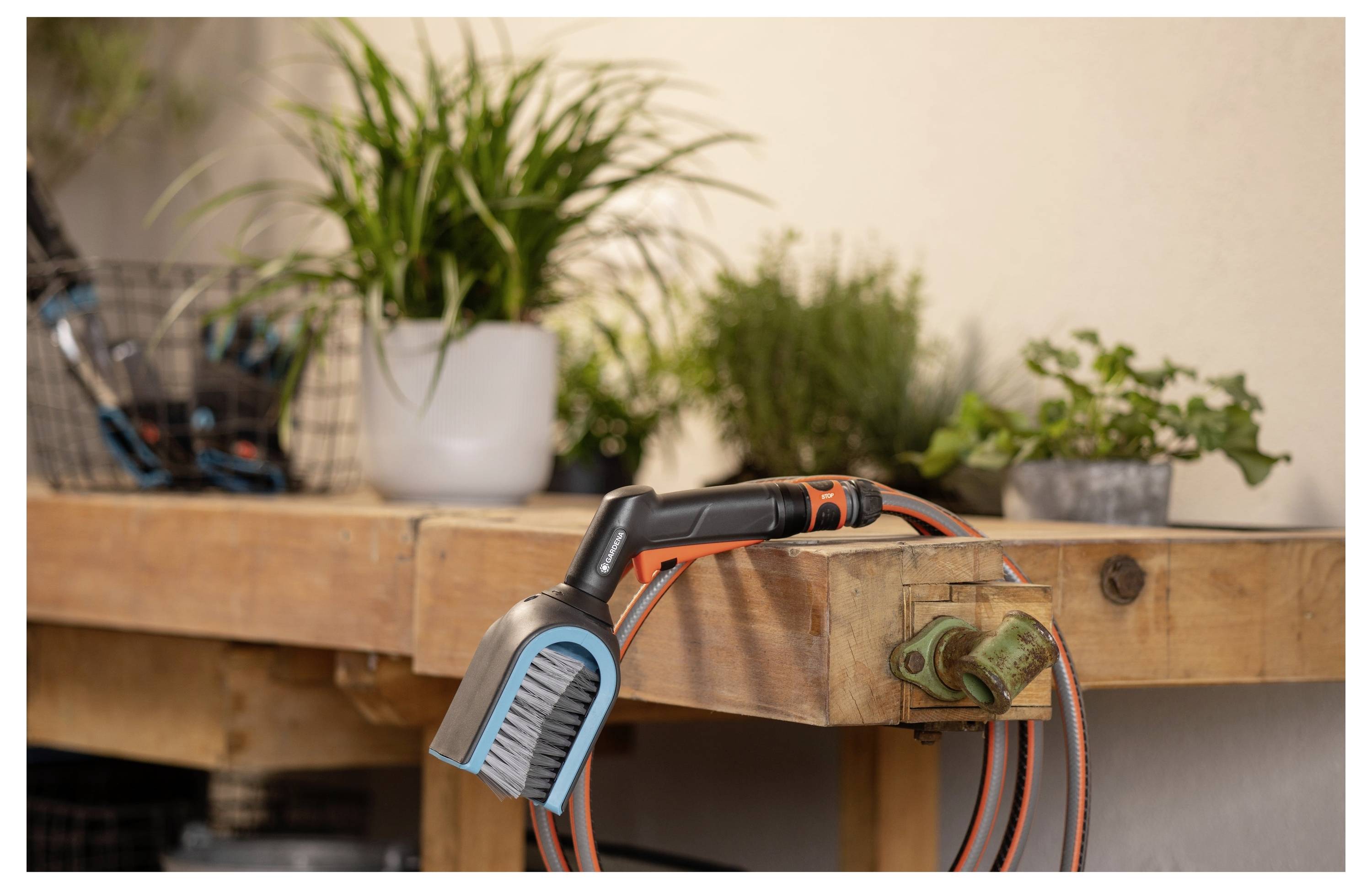 Wąż z wyspecjalizowaną dyszą spoczywa na drewnianym warsztacie, otoczony doniczkowanymi roślinami, co sugeruje ogrodniczy zestaw.