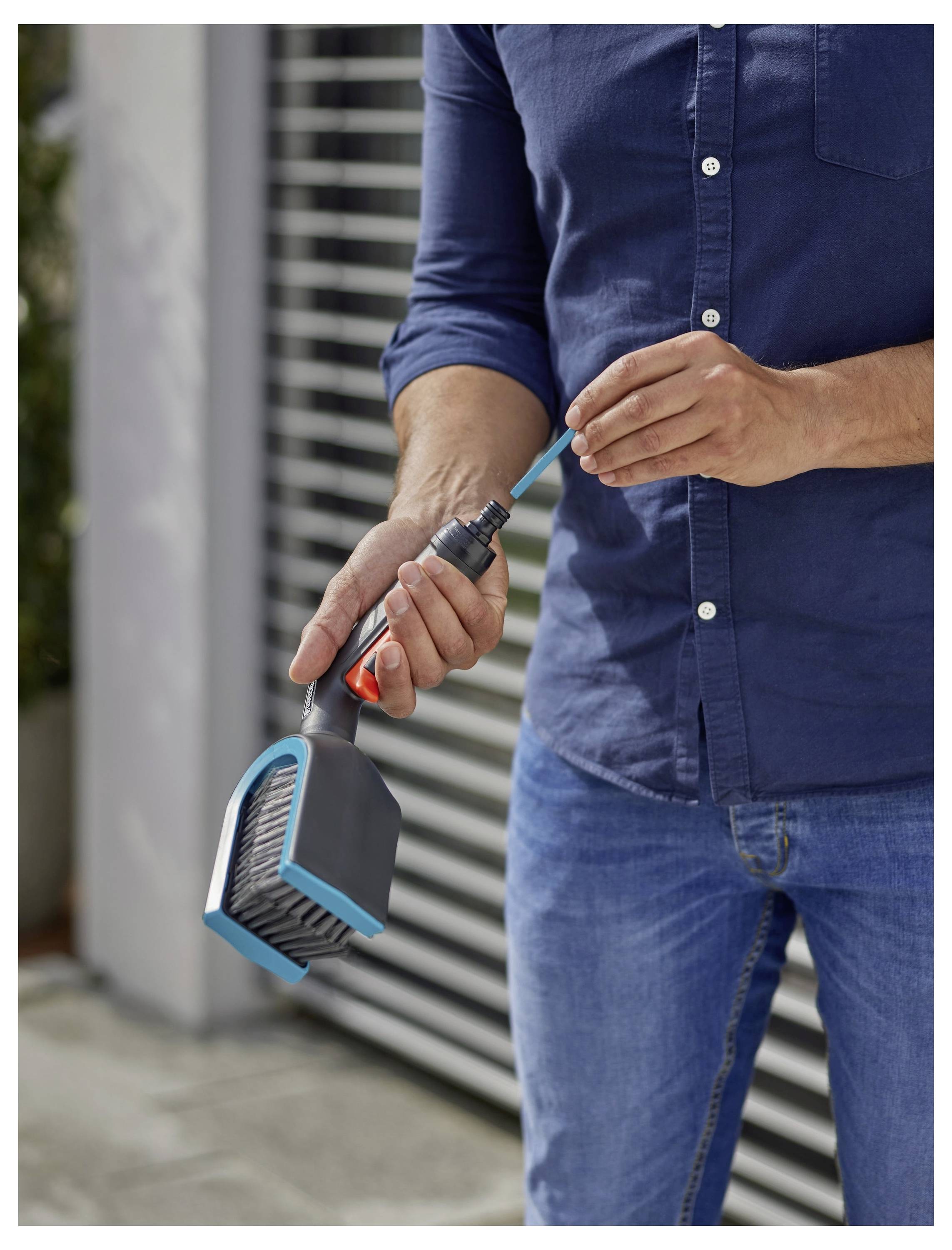 Osoba w niebieskiej koszuli podłącza narzędzie szczotki do węża ogrodowego w celu czyszczenia, stojąc na zewnątrz przed nowoczesnym budynkiem.