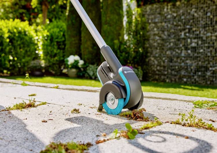 Ręczne urządzenie z szczotką usuwa chwasty z płyty tarasowej. W tle widoczne są zielone krzewy.