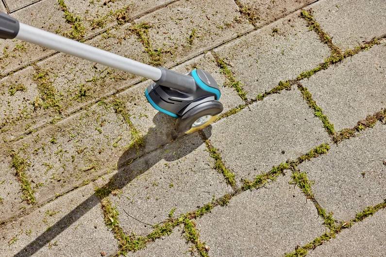 Osoba używa narzędzia ogrodowego do usuwania chwastów między kostką brukową. Zdjęcie przedstawia zbliżenie urządzenia podczas pracy.