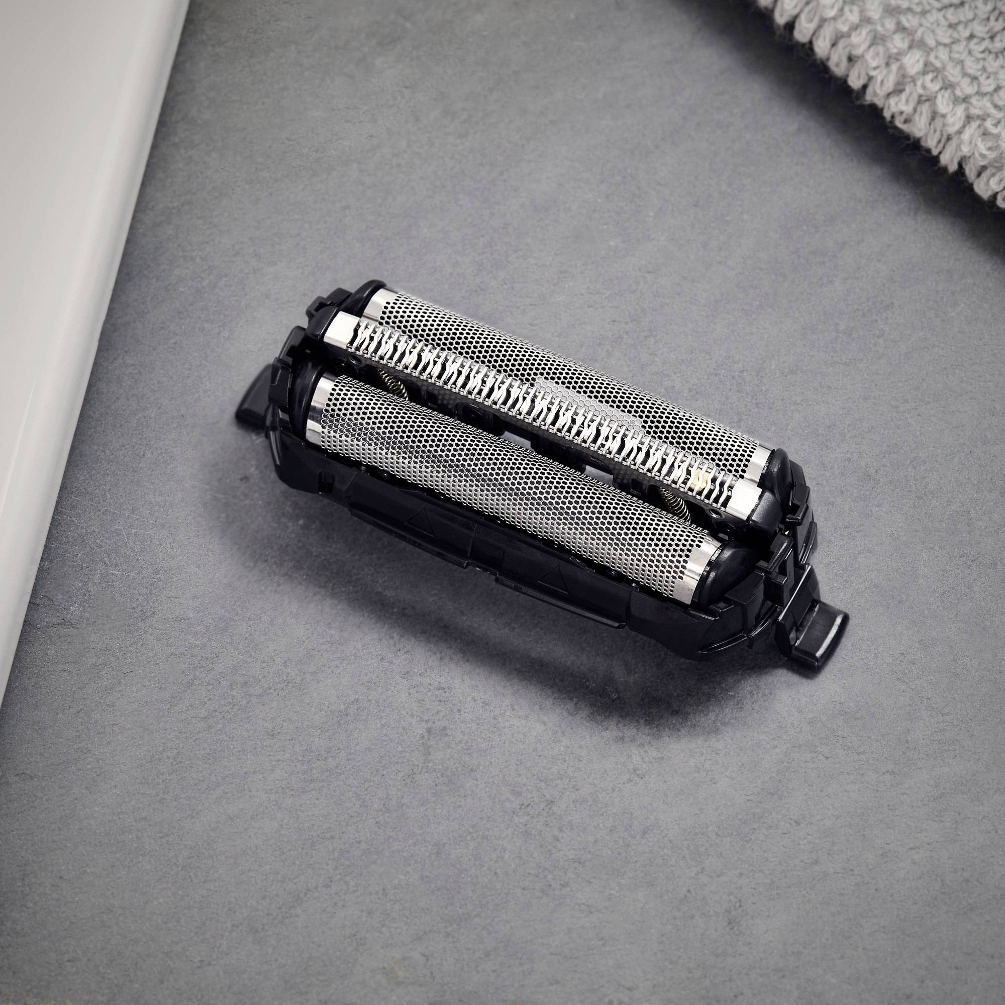 Głowica golarki z dwoma obracającymi się ostrzami metalowymi na ciemnym tle, obok szarej szmatki, symbolizuje pielęgnację ciała.