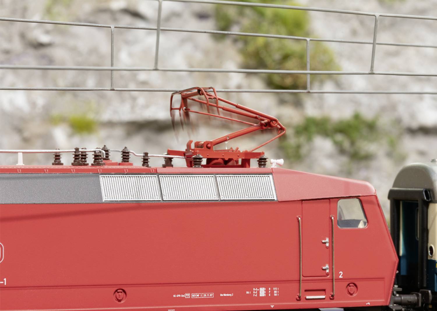 Czerwona lokomotywa z podniesionymi pantografami pod siecią trakcyjną. Tło rozmyte, z widocznymi skałami i roślinnością.