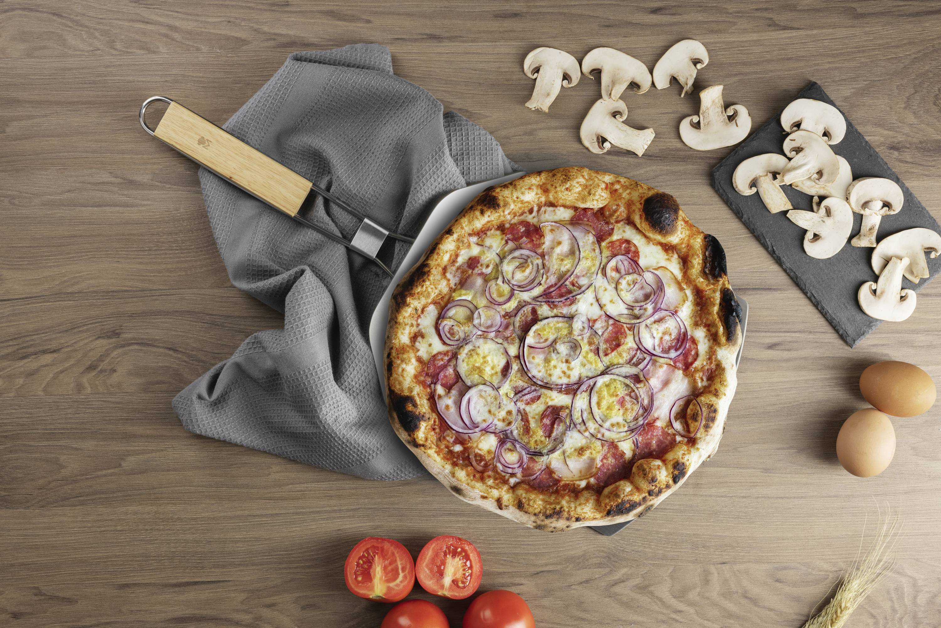 Pizza z serem, cebulą i kiełbasą na łopacie do pizzy, obok pokrojonych pieczarek, jajek i pomidorów na drewnianym stole.
