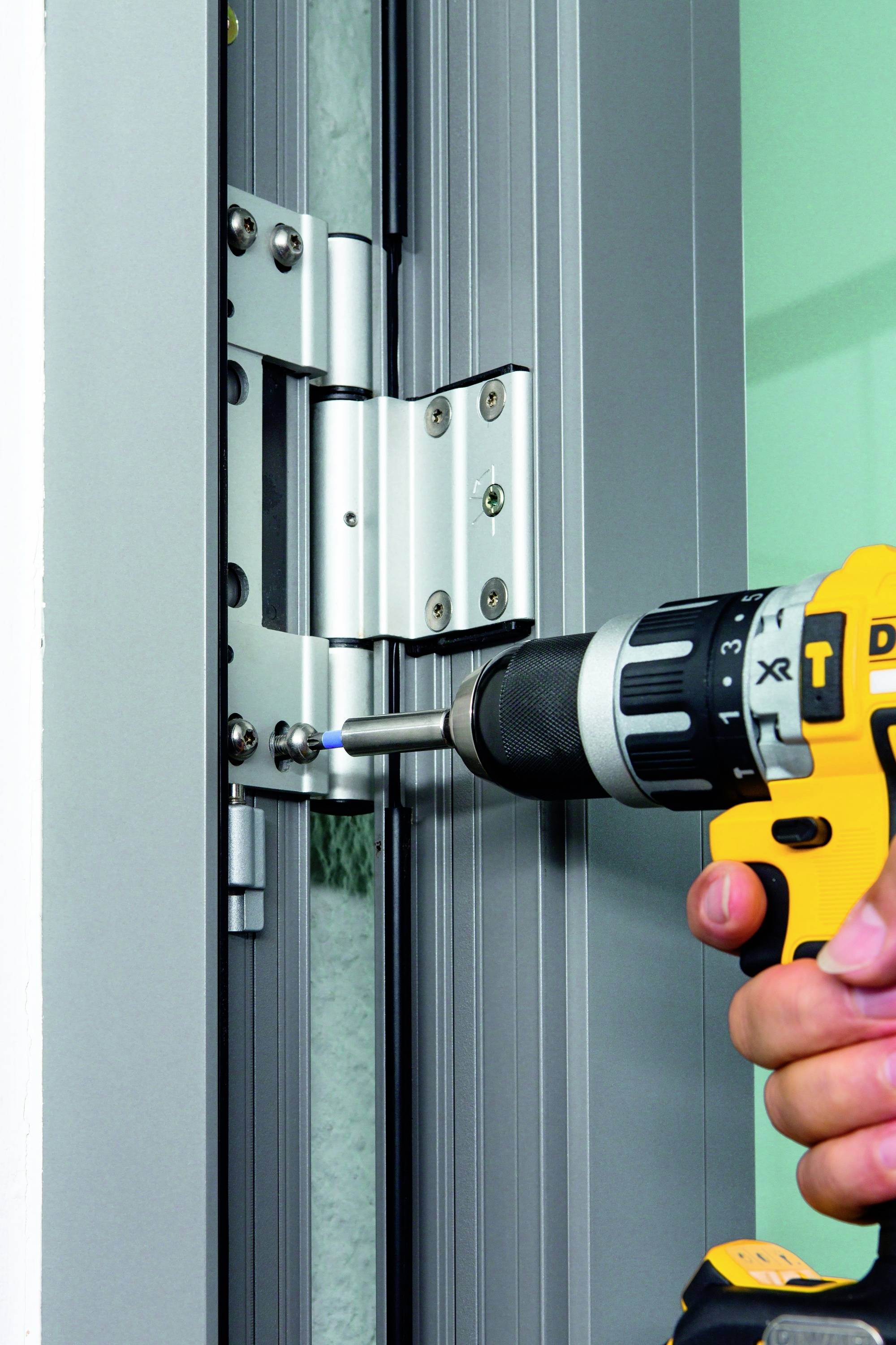Osoba mocuje zawias do drzwi za pomocą akumulatorowego wkrętarki. Zawias jest srebrny, a wkrętarka żółto-czarna.