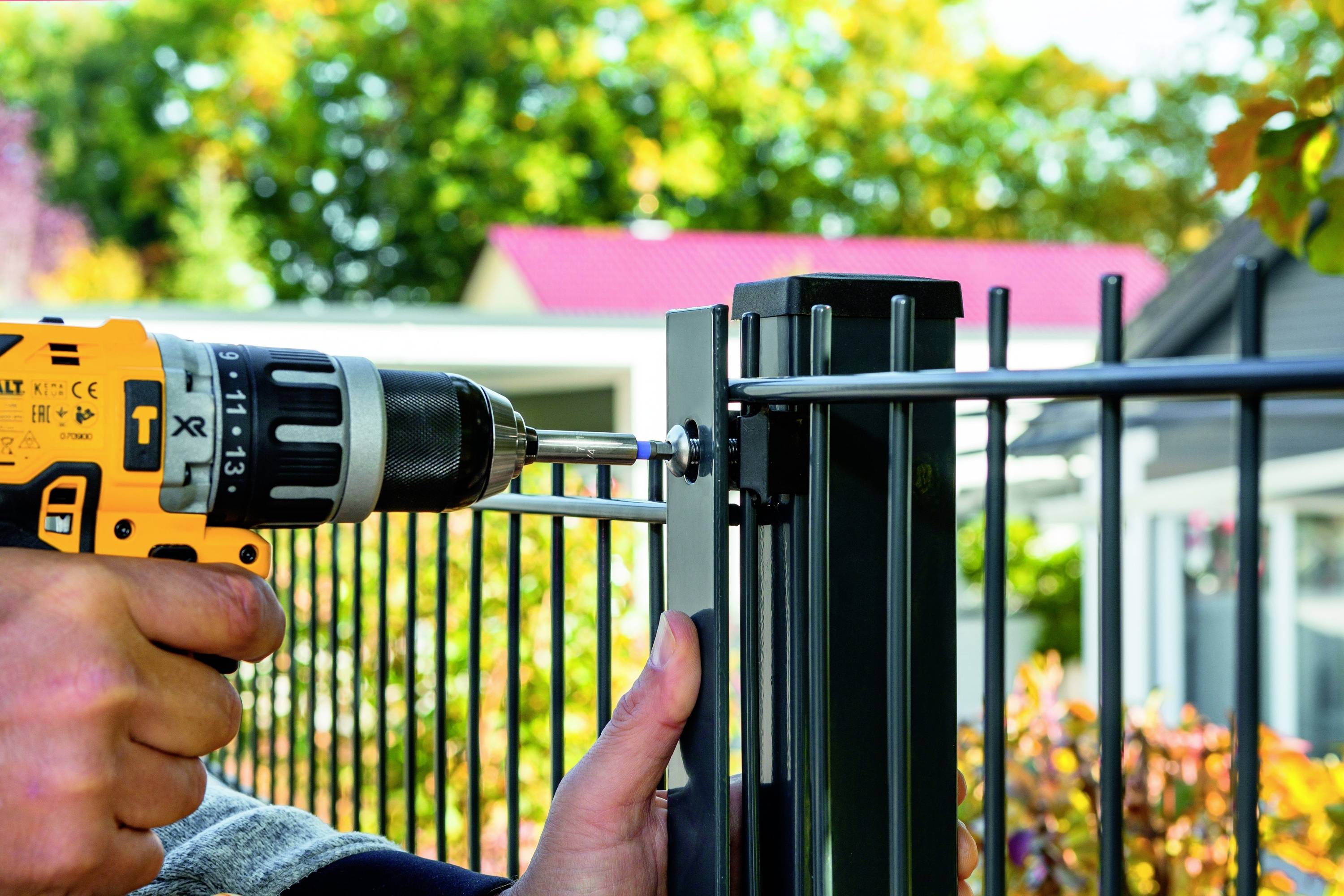 Osoba mocuje element metalowego ogrodzenia wiertarką do metalowego słupa. W tle: domy i jesienne drzewa.
