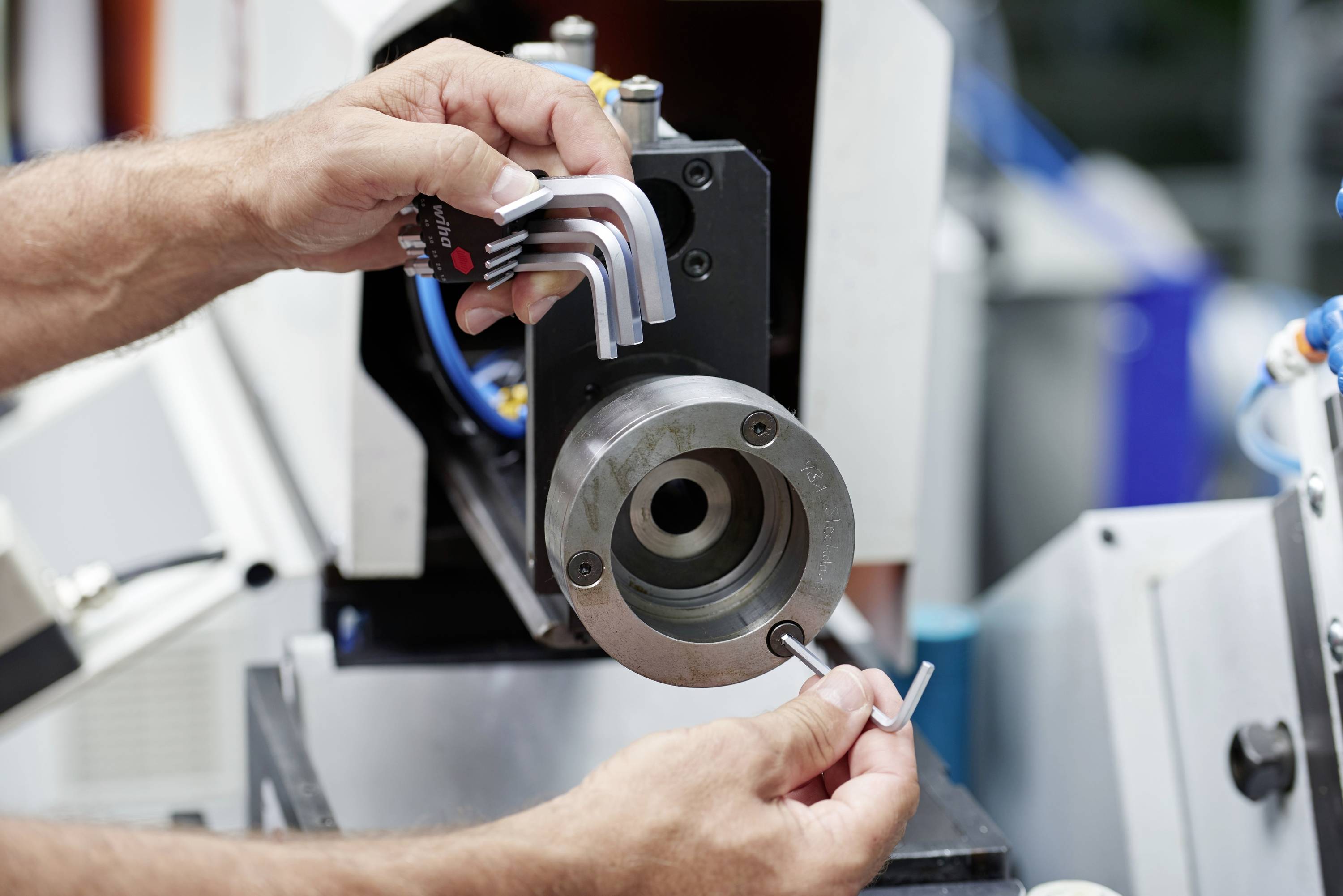 Dwie ręce regulują maszynę za pomocą zestawu kluczy imbusowych trzymanych przed metalowym cylindrem, prawdopodobnie w warsztacie.