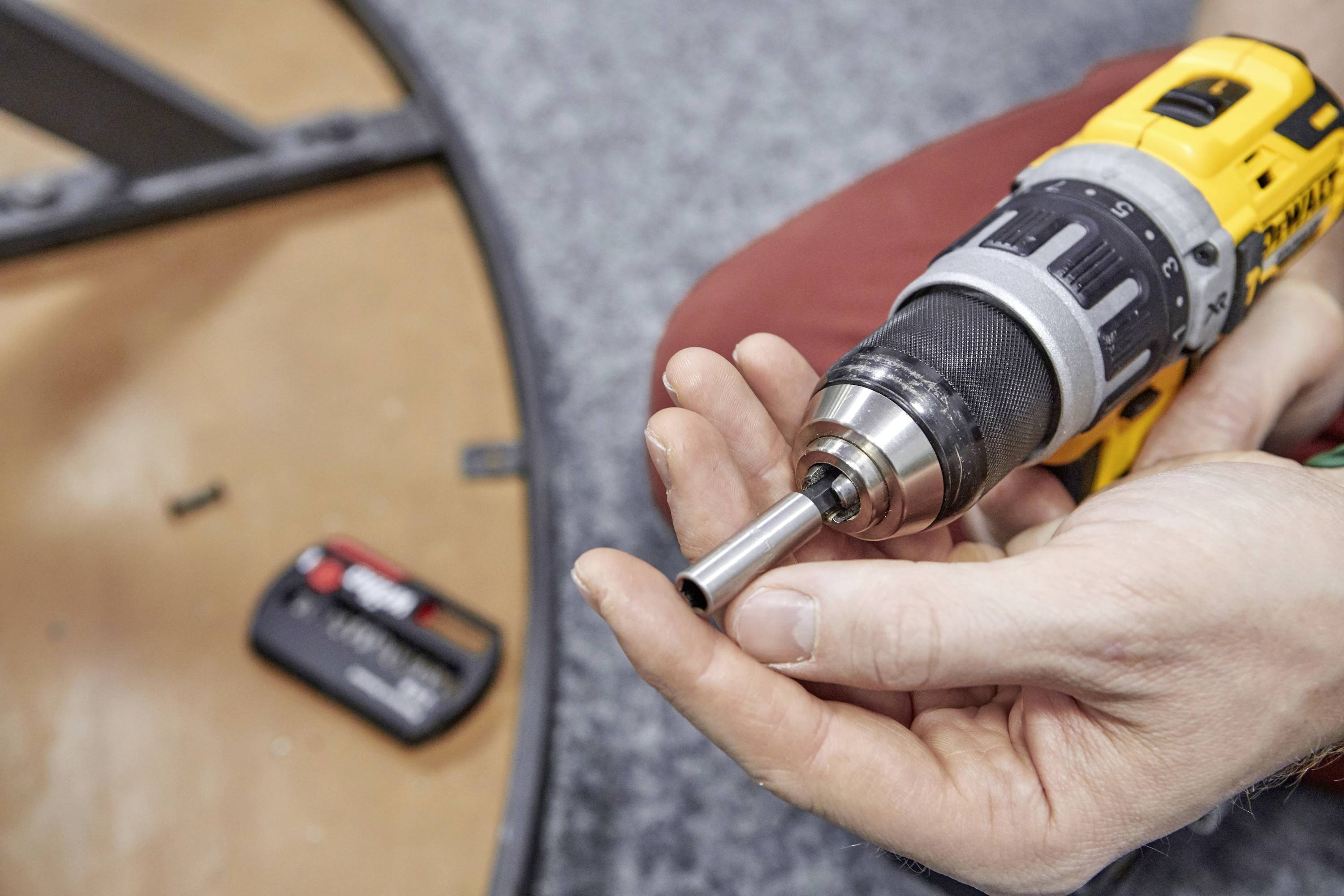 Jedna ręka trzyma akumulatorową wkrętarkę z włożonym wiertłem. Na tle widać poziomicę leżącą na dywanie.