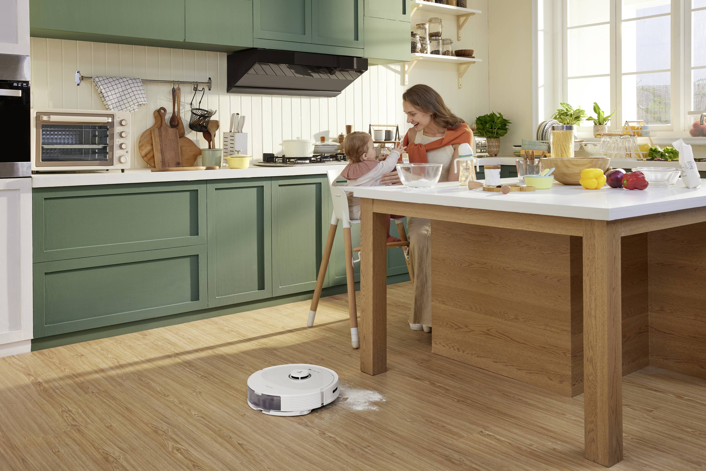 Kobieta karmi dziecko w nowoczesnej kuchni; robot sprzątający zbiera rozsypane na podłodze mąki.