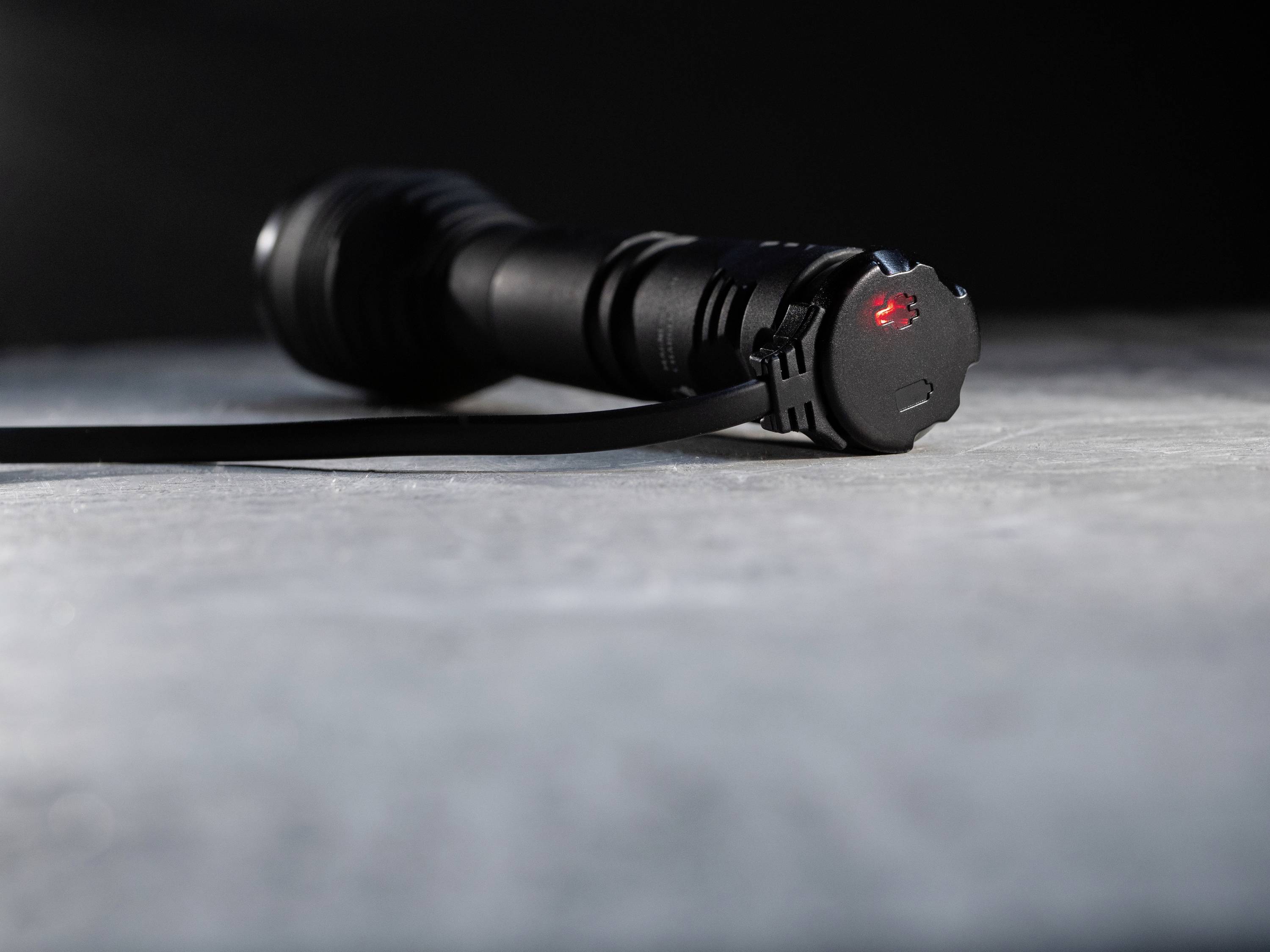 Czarna latarka leży poziomo na szarej powierzchni. Na jej końcu słabo świeci czerwone światło.
