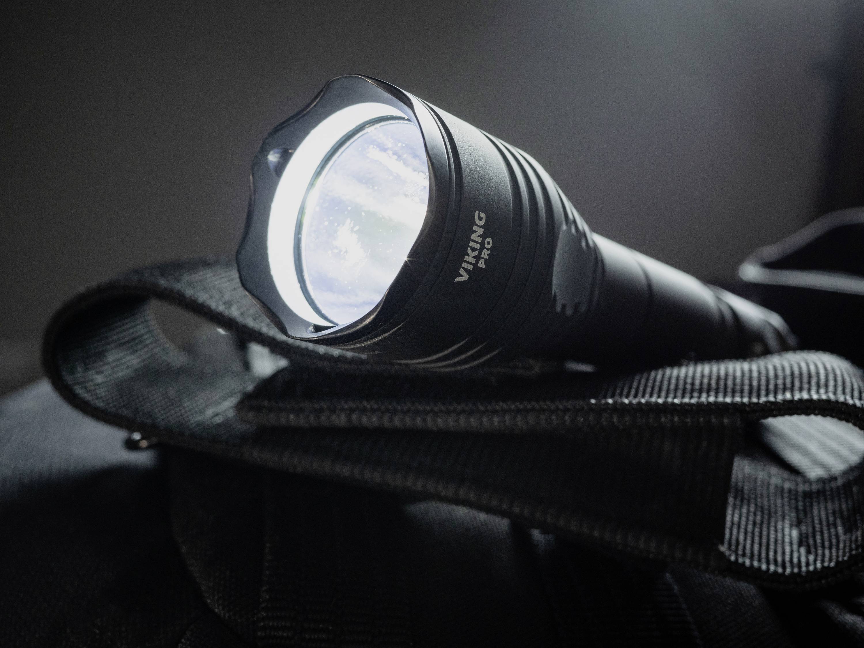 Włączona latarka leży na czarnej torbie. Świeci jasno i wyróżnia się na ciemnym tle.