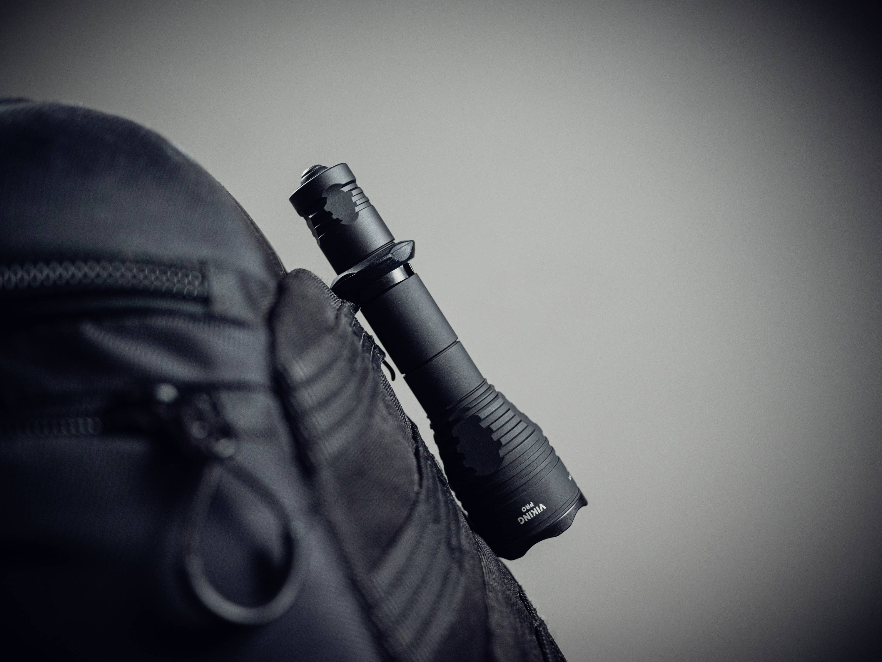 Czarna latarka jest włożona z boku do kieszeni czarnego plecaka. Tło jest rozmyte i ciemnoszare.