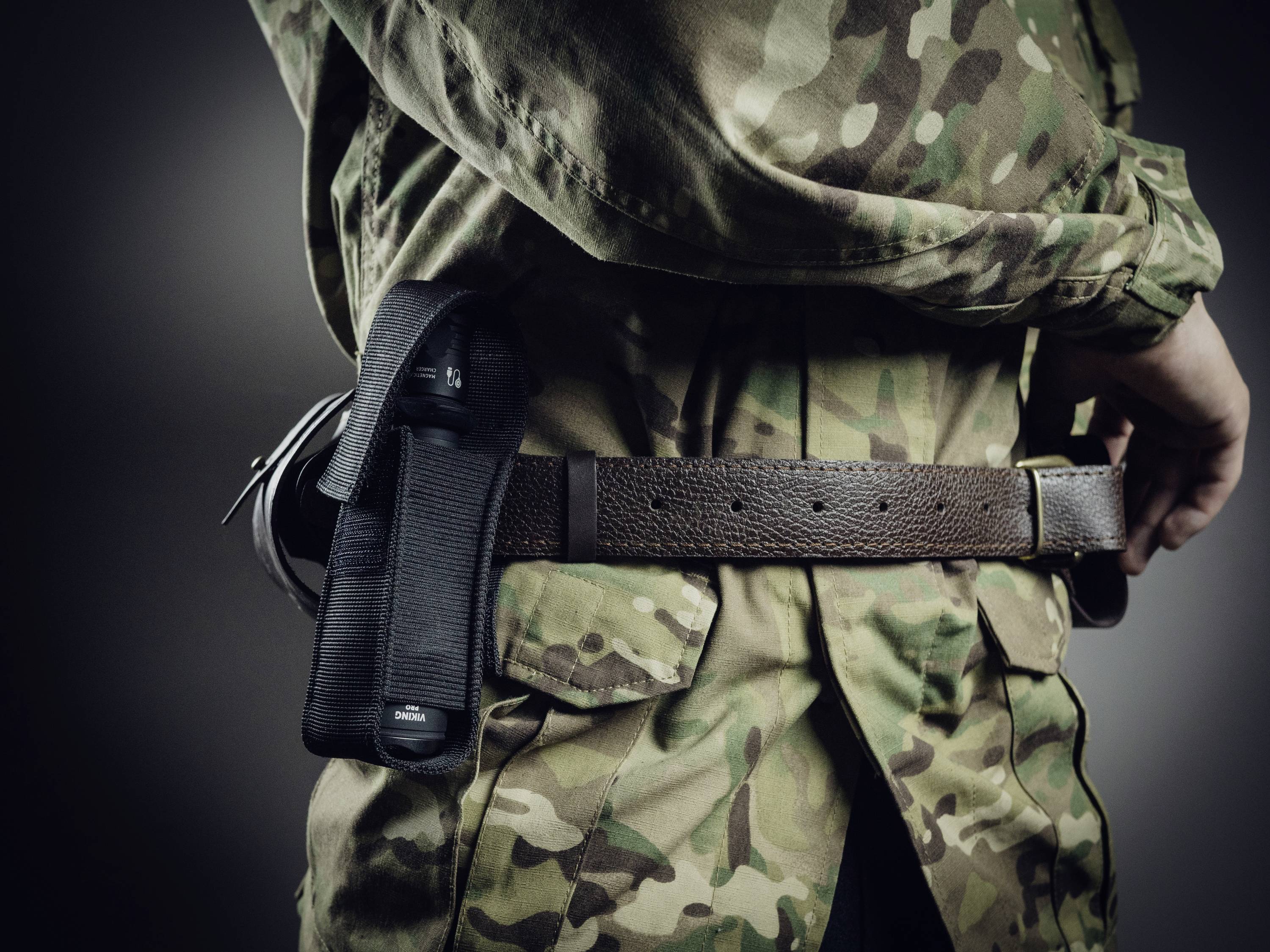 Osoba w mundurze maskującym nosi broń krótką w kaburze przy pasie, sfotografowana z tyłu z boku.