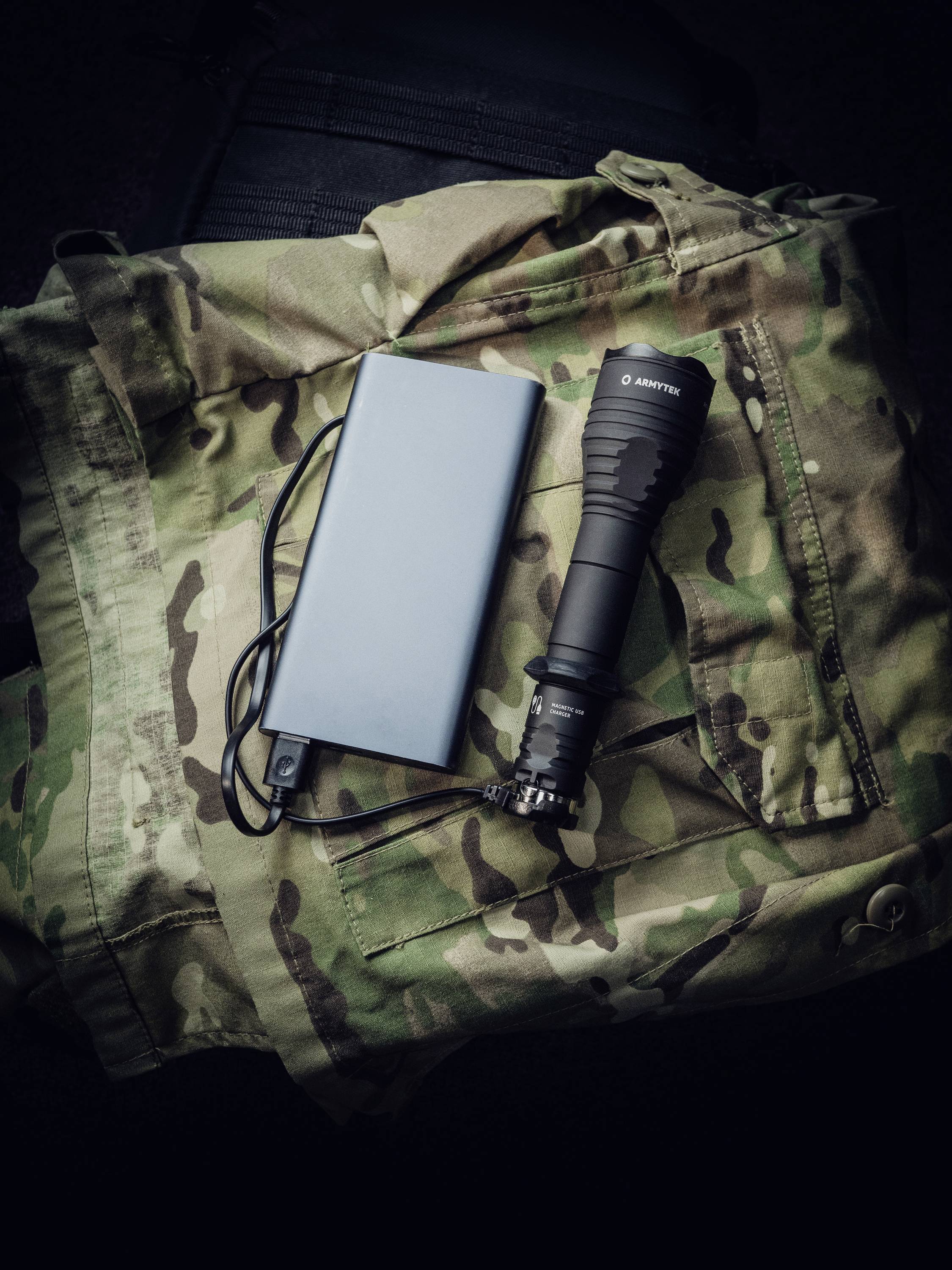 Latarka, powerbank i kabel leżą na kamuflażowej odzieży.