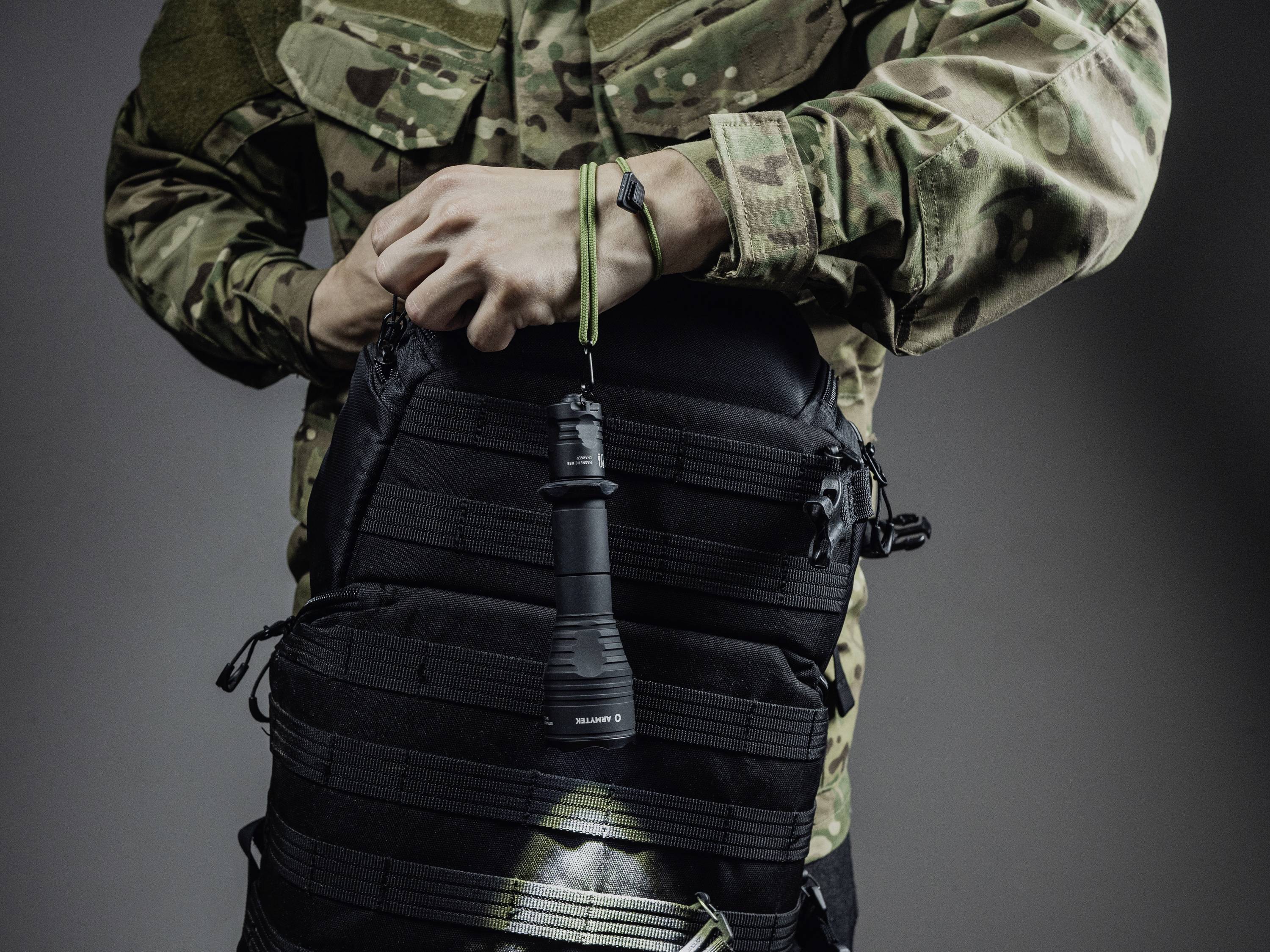 Człowiek w kamuflażu trzyma czarną latarkę przymocowaną do czarnego plecaka.