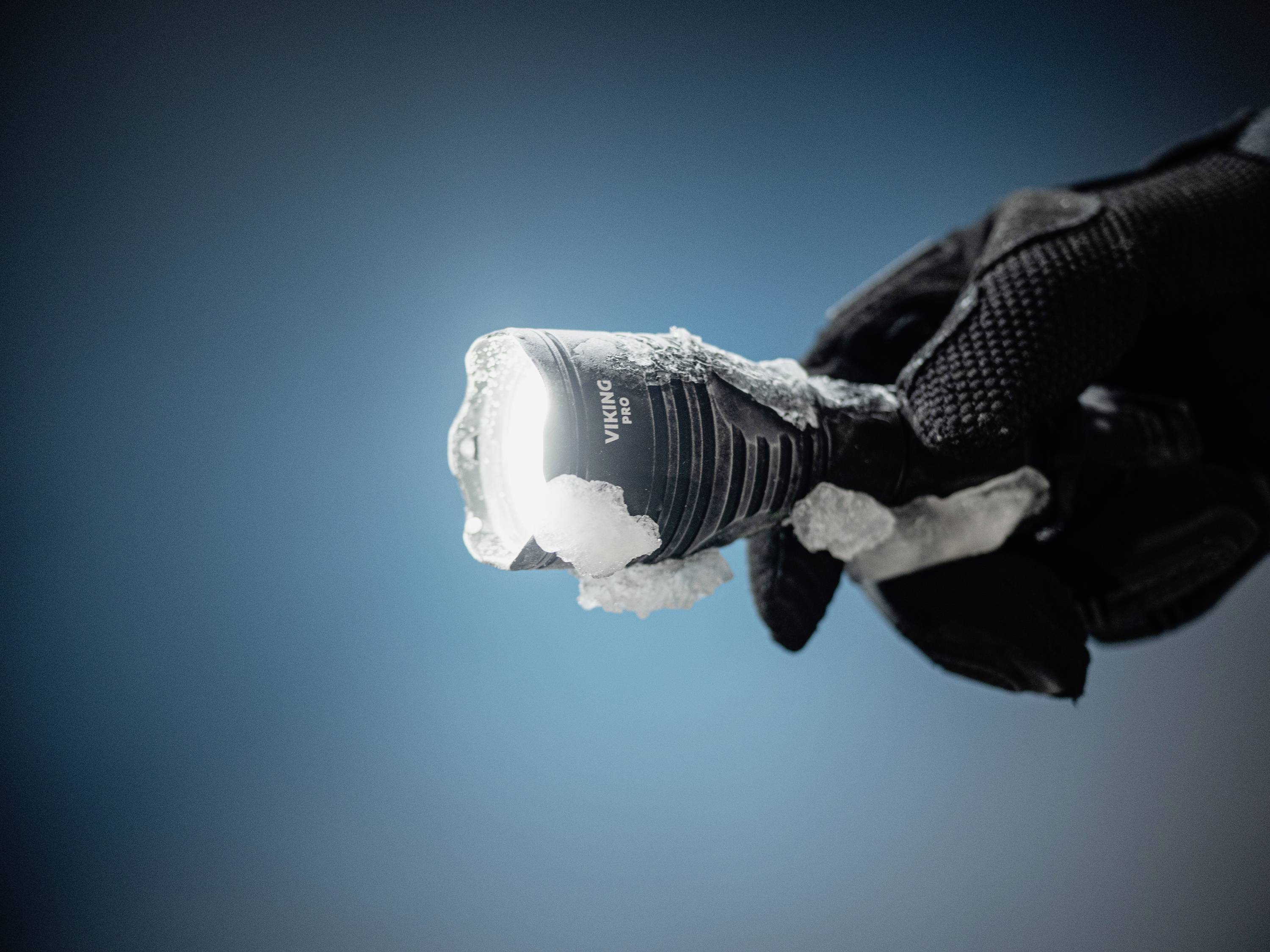 Rękawiczkowana dłoń trzyma ośnieżoną, świecącą latarkę na niebieskim tle.