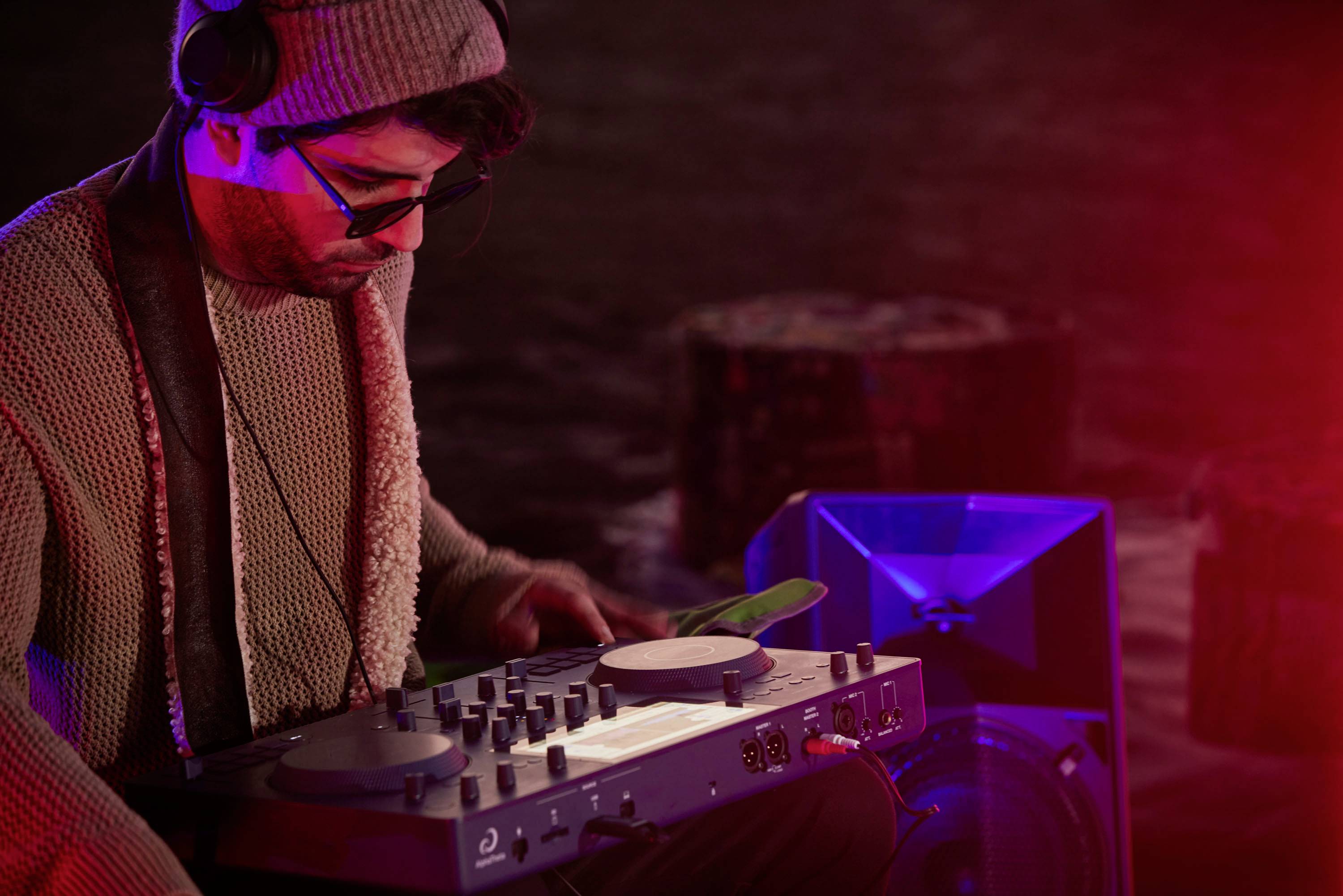 DJ z zestawem słuchawkowym miesza muzykę na mikserze w klimatycznym pomieszczeniu oświetlonym czerwonym światłem. W tle widać wodę i światła.