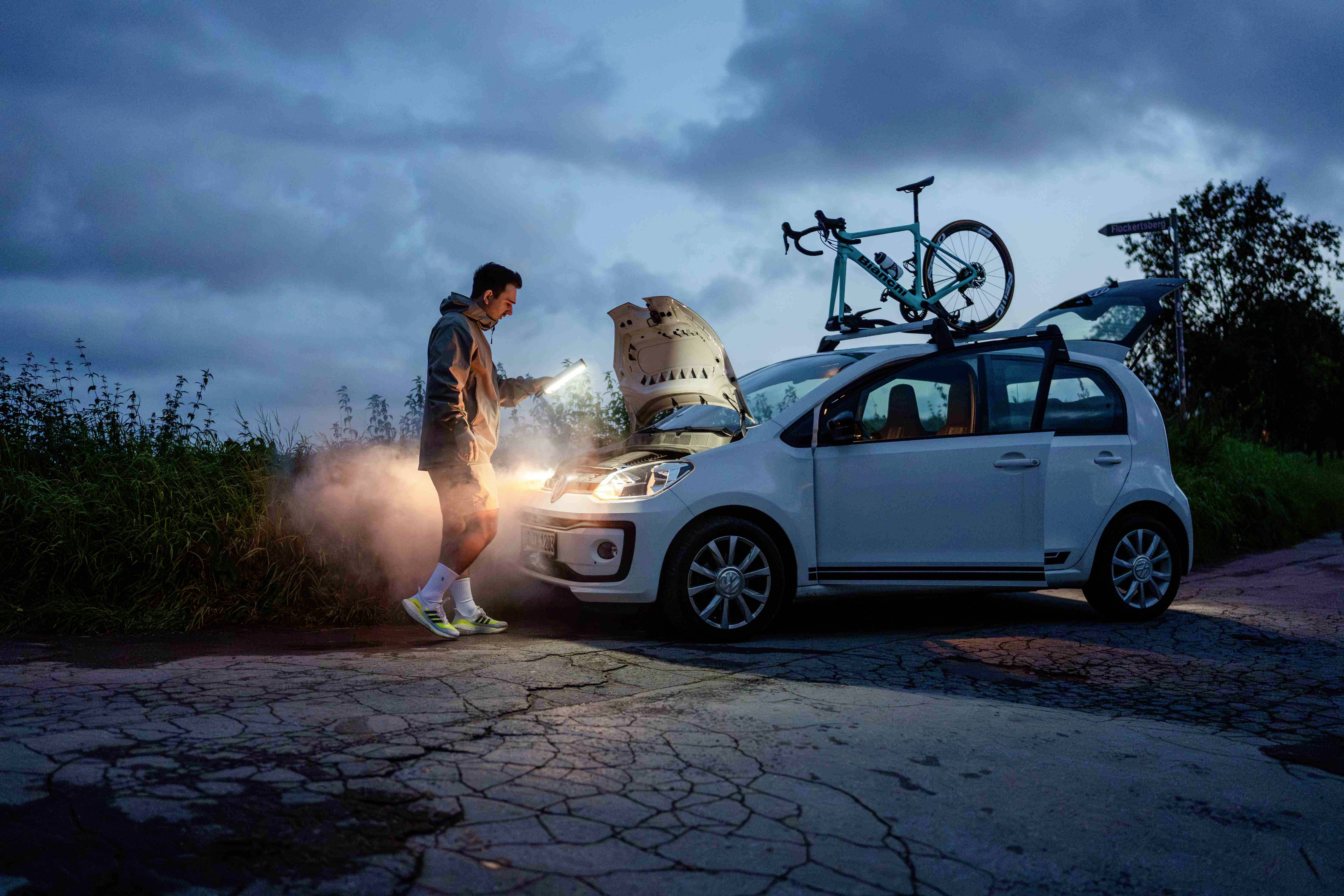 Mężczyzna z latarką stoi przy białym samochodzie z otwartą maską na poboczu drogi, a na dachu zamontowany jest rower.