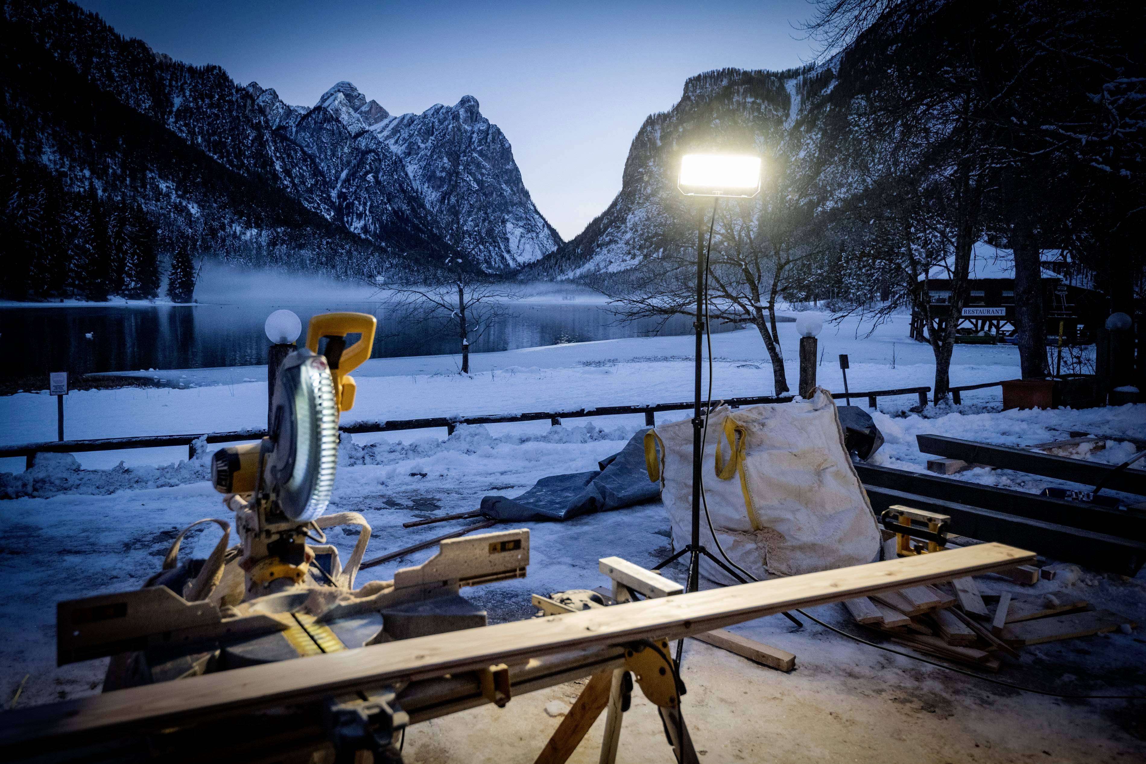 Piła na stole przed ośnieżonym jeziorem górskim o zmierzchu, oświetlona jasnym reflektorem budowlanym. Majestatyczny krajobraz górski w tle.