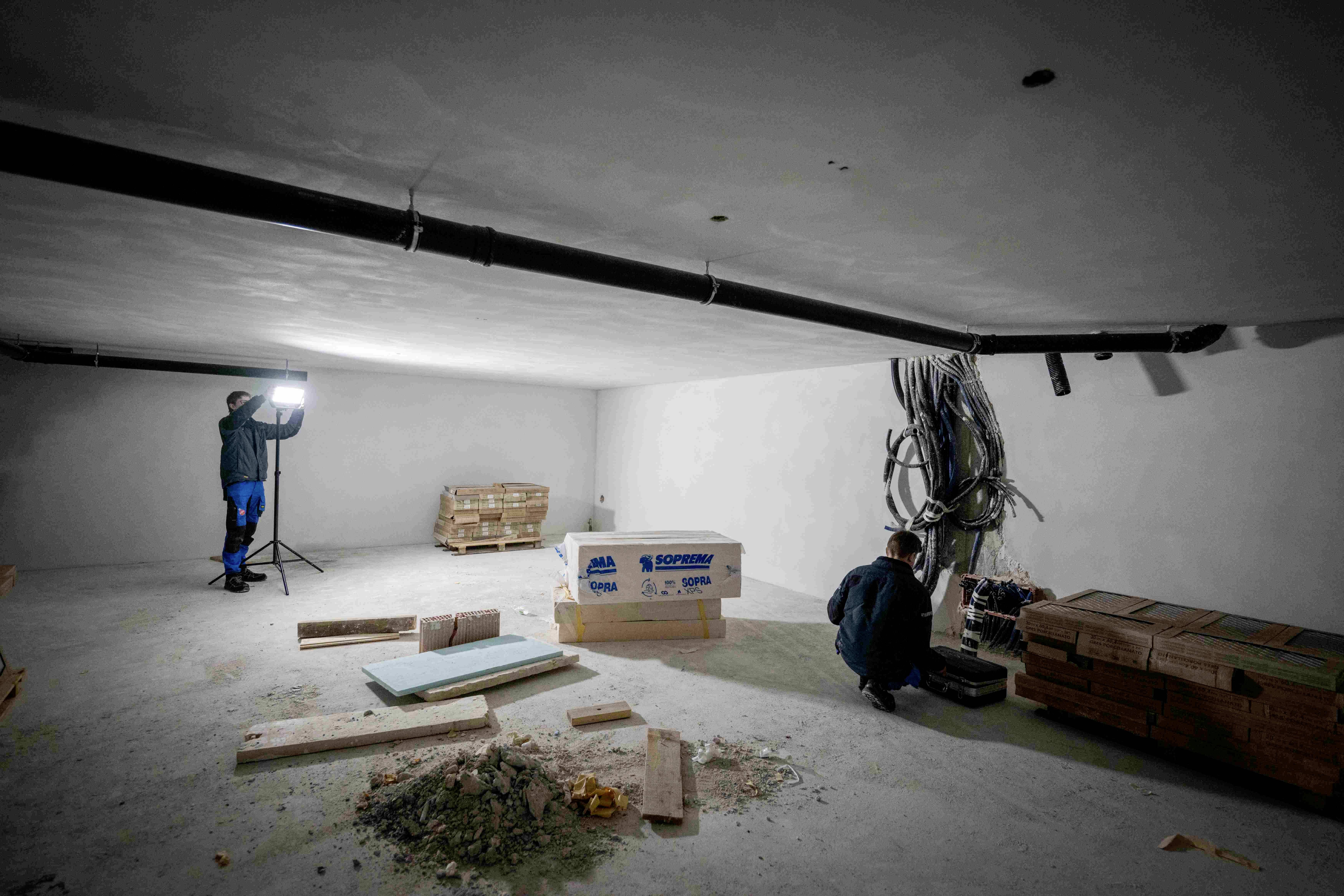 Pusta plac budowy w piwnicy, dwóch robotników, materiały i narzędzia rozrzucone dookoła. Jeden z robotników stoi przed lampą.