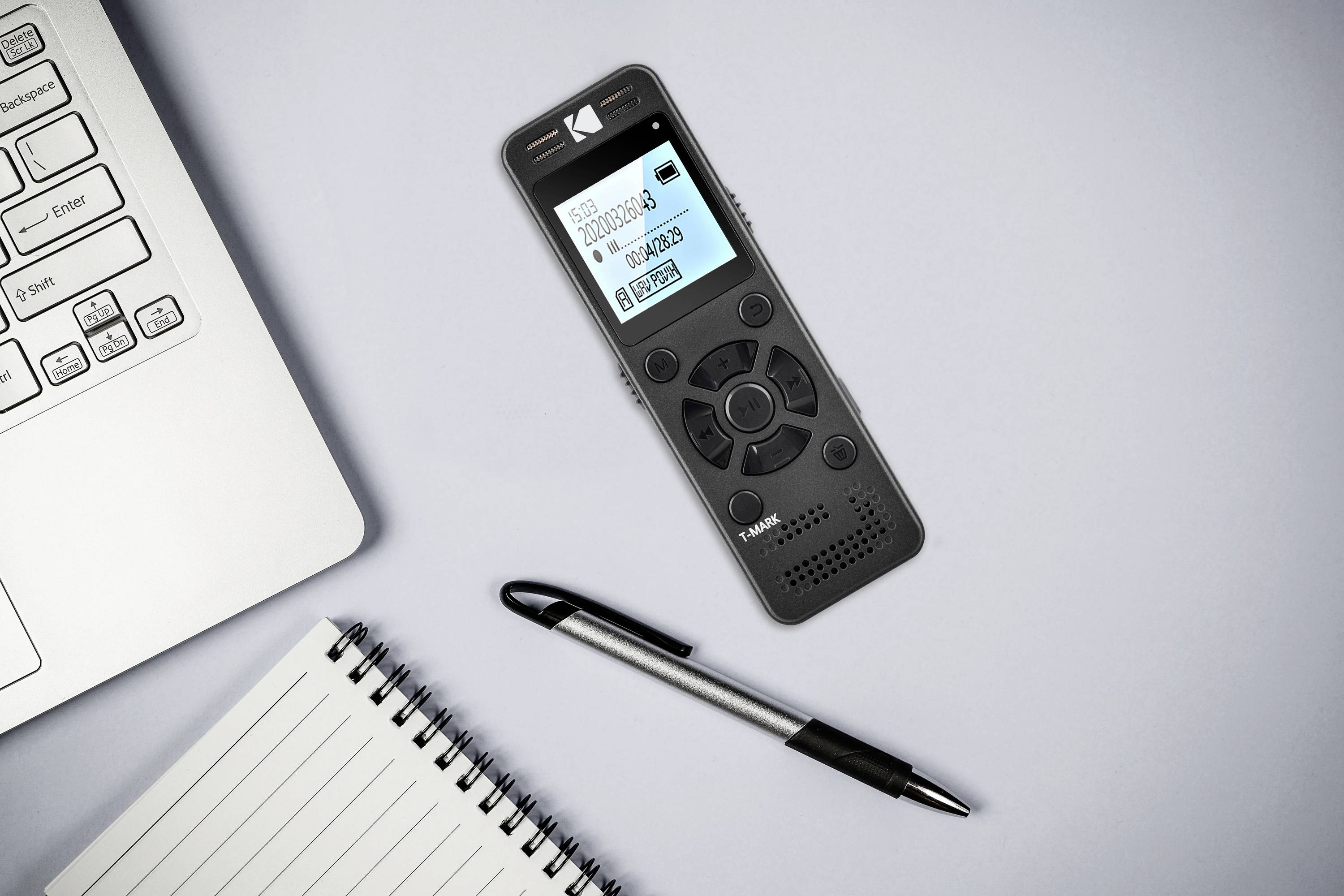 Dyktafon z cyfrowym wyświetlaczem leży obok laptopa, długopisu i notesu na stole. Urządzenie wyświetla informacje o nagraniu.