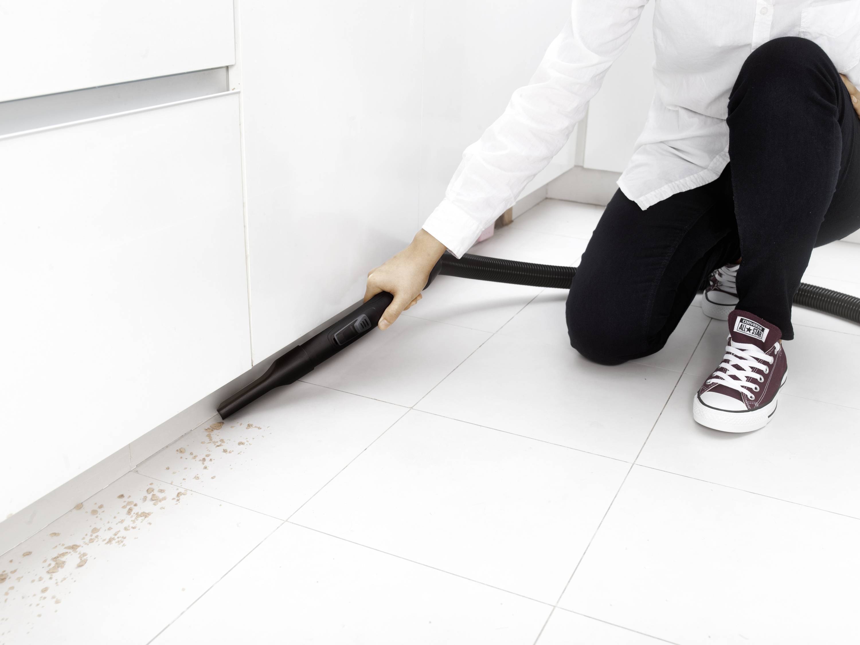 Osoba ubrana w białą koszulę i ciemne spodnie odkurza okruchy z białej podłogi kuchennej, klęcząc.