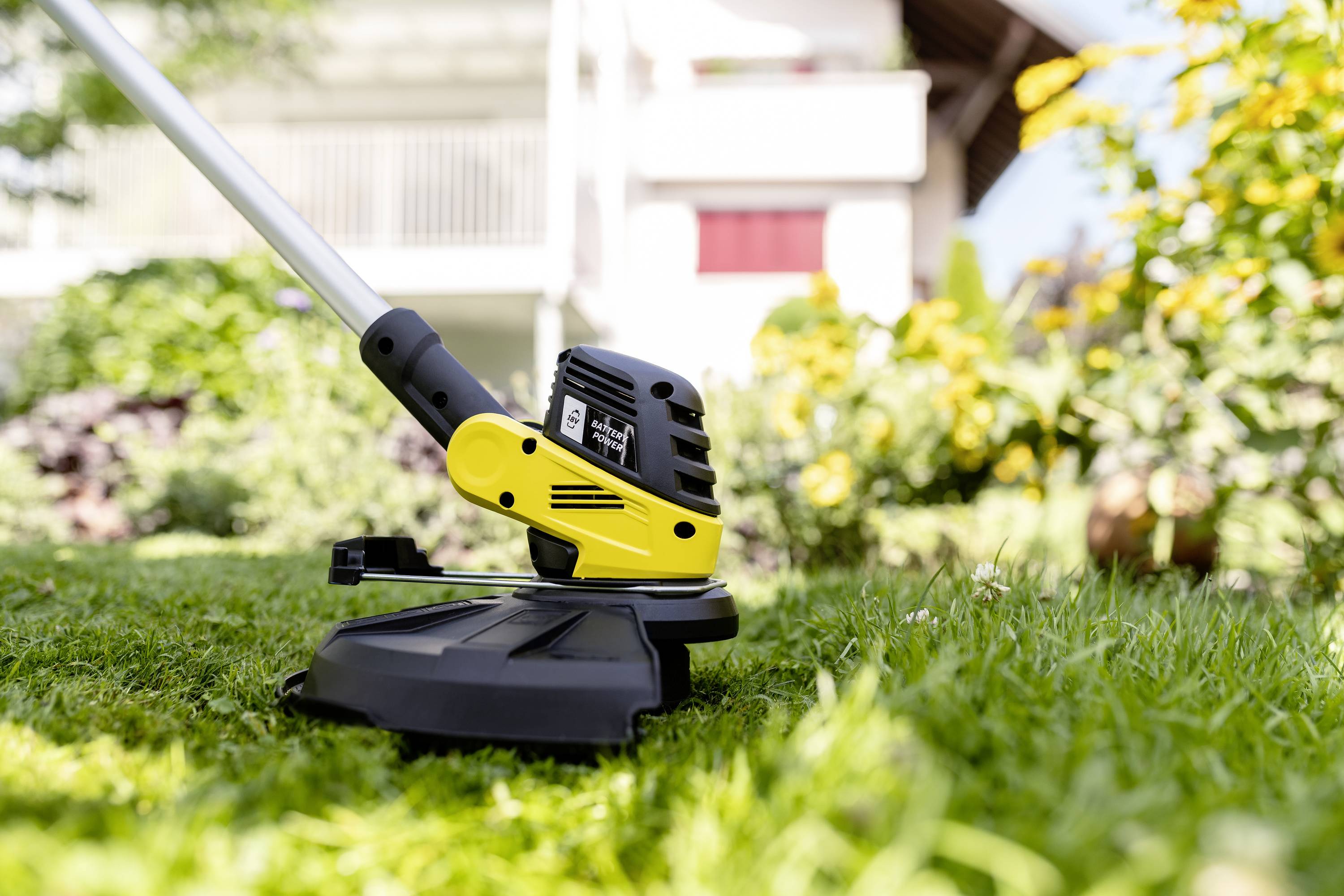 Trymer do trawy przycina trawę w ogrodzie. W tle niewyraźnie widać kwiaty i budynek.