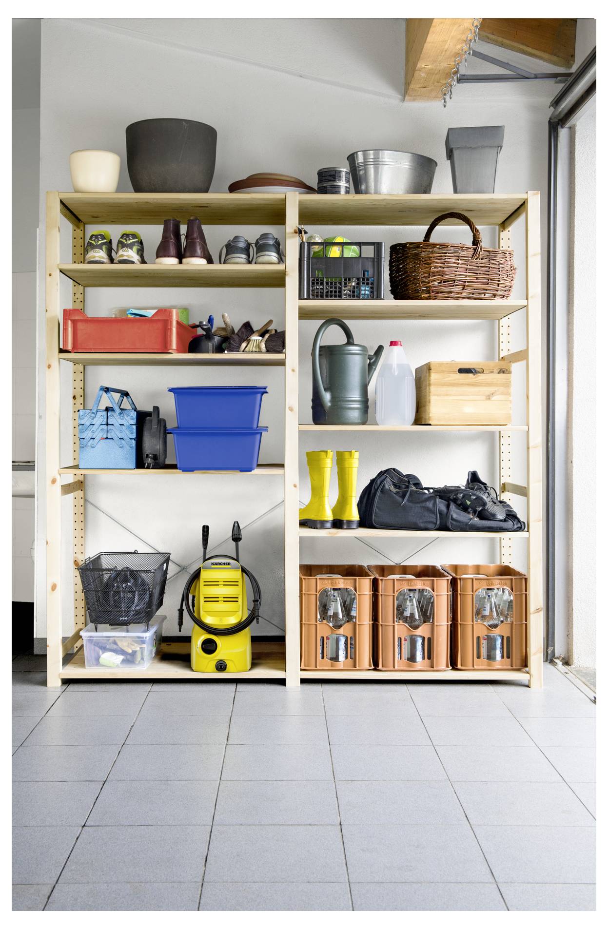 Drewniane półki w garażu przechowują narzędzia ogrodnicze, buty, pojemniki magazynowe i środki czystości. Widoczne są jasno żółte buty i odkurzacz.